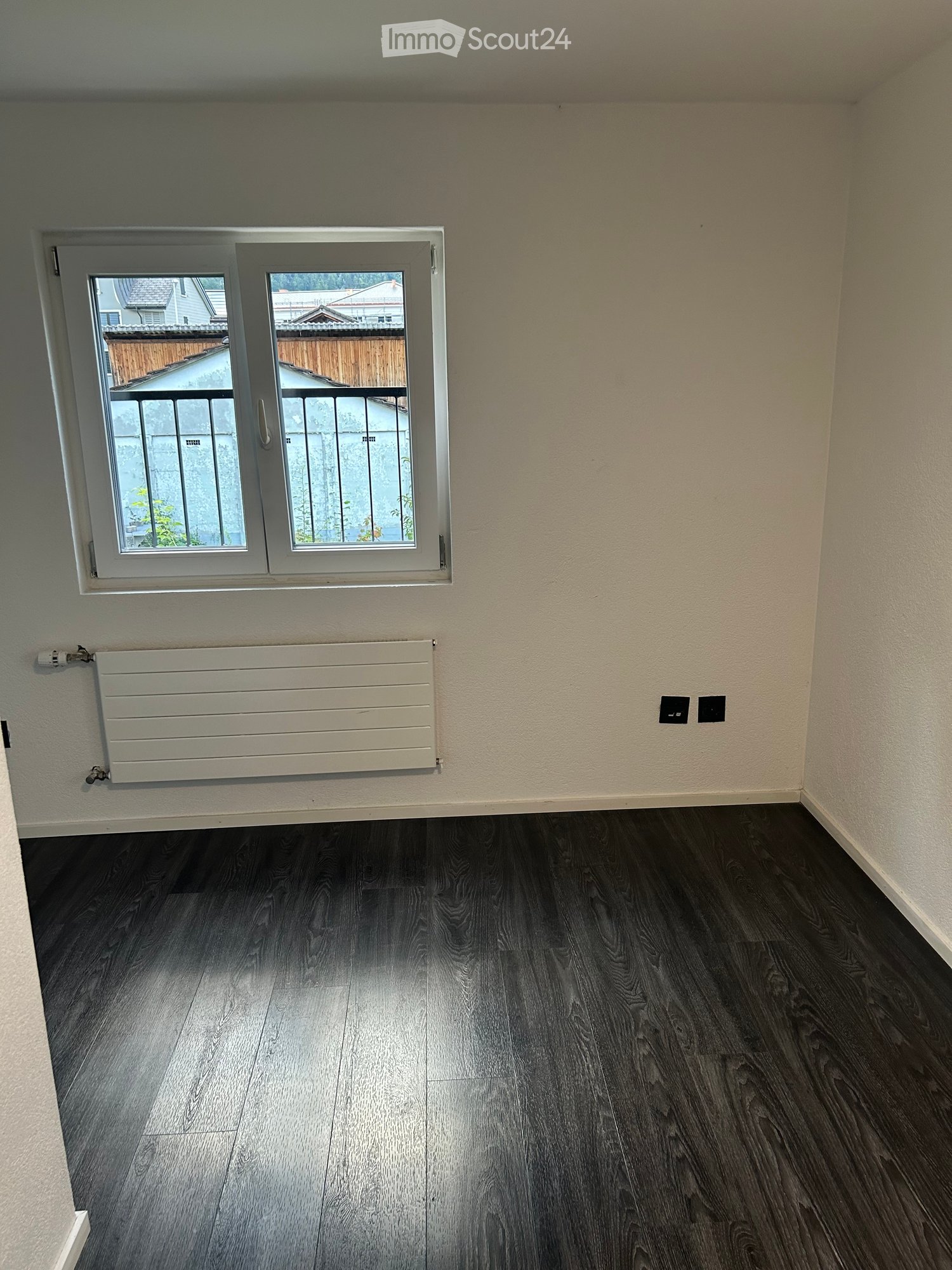 Empty room with dark wooden flooring, white walls, a window with a view of a balcony, and a radiator below the window.