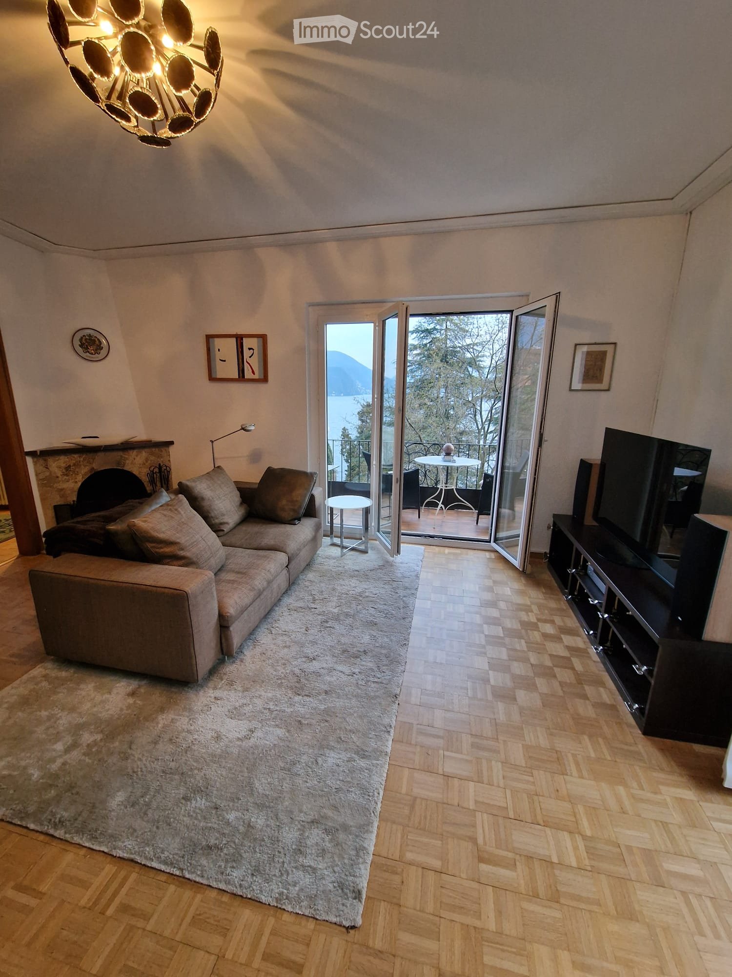 Modern living room with a large sectional sofa, coffee table, TV, fireplace, and a view of the lake through large glass doors leading to a balcony.