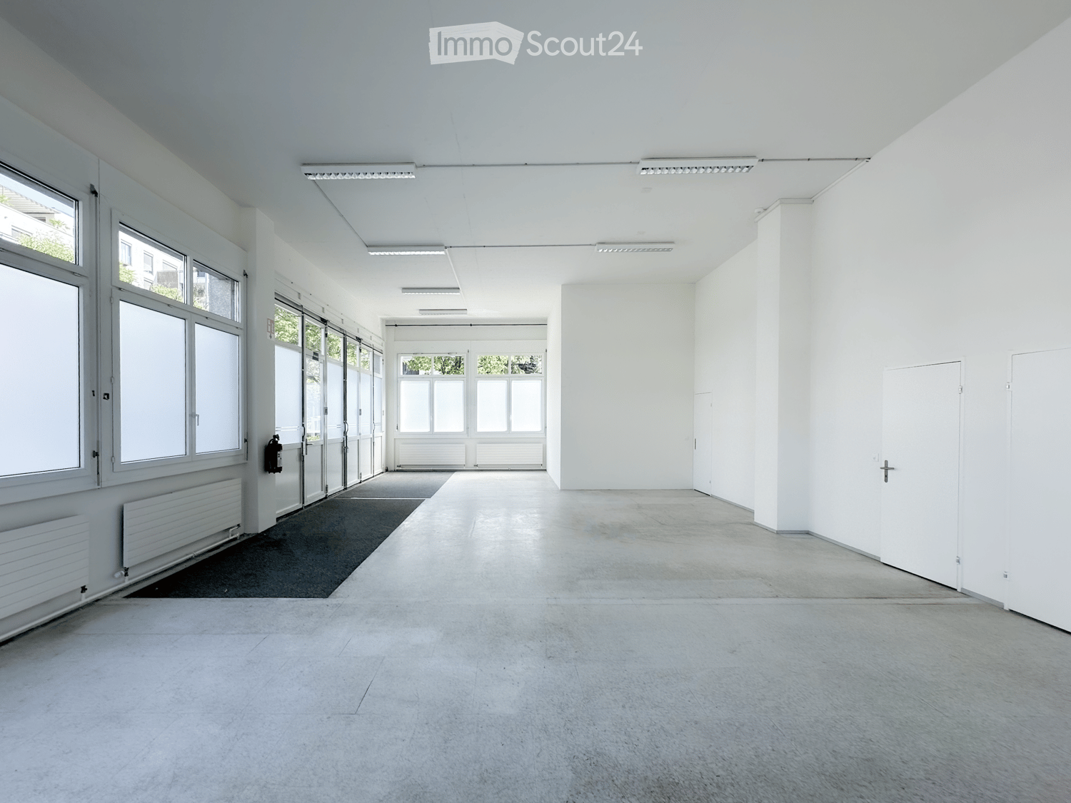 Empty room with white walls, large windows, and a concrete floor. No furniture or equipment is present.
