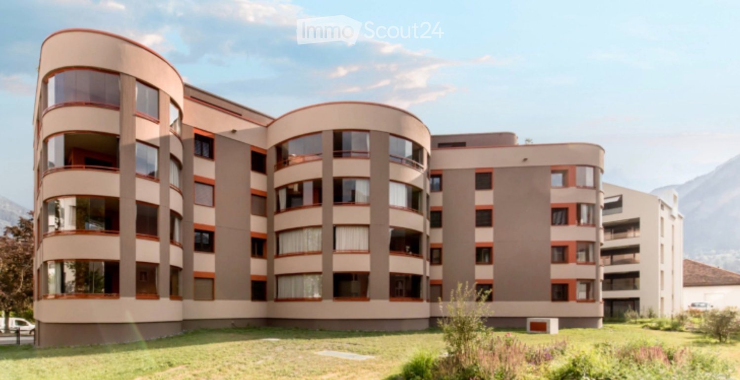 Modern multi-story building with curved sections, large windows, and balconies, set against a scenic mountain backdrop.