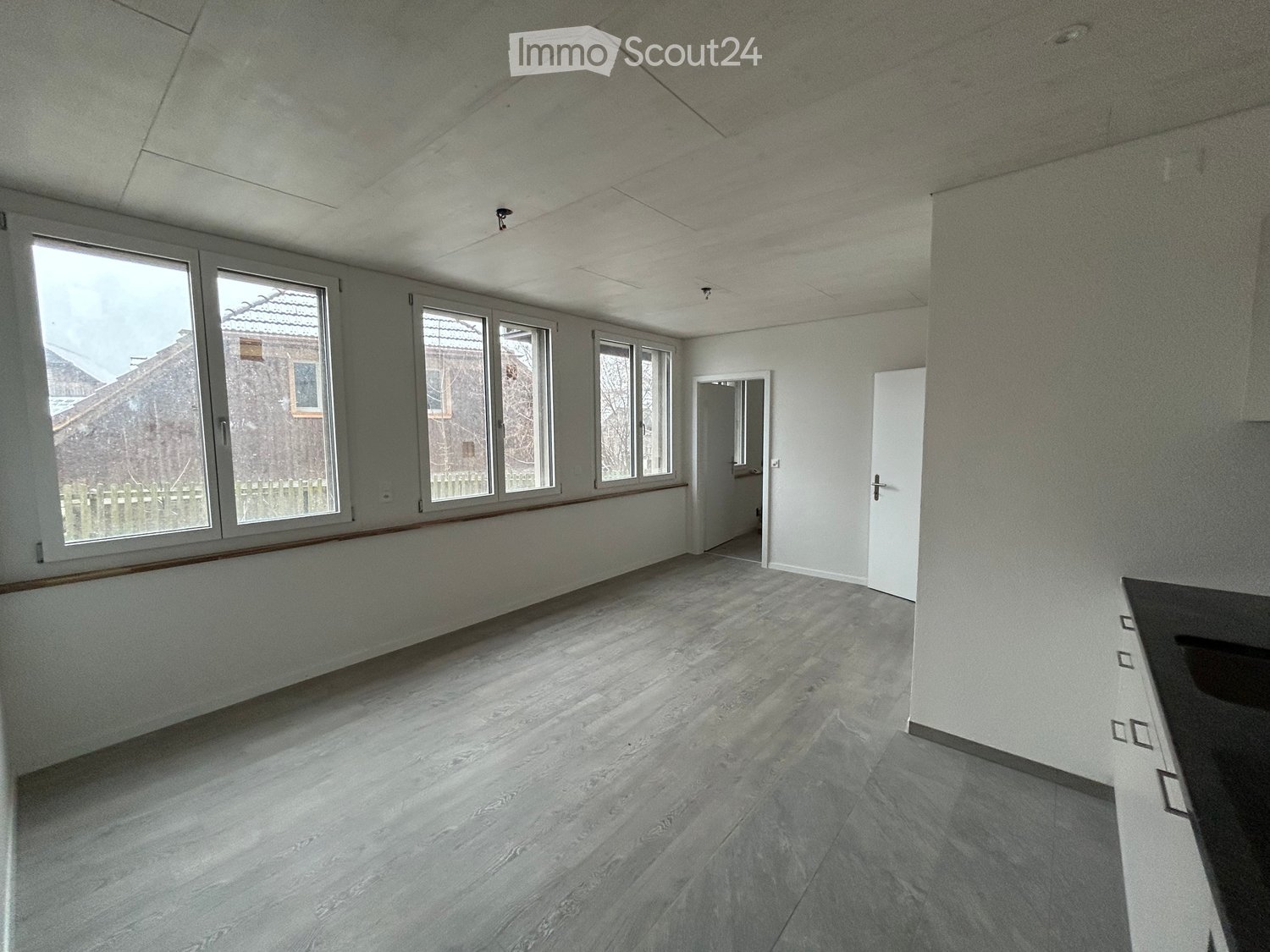 Empty room with light-colored walls, wooden flooring, and large windows allowing natural light. The space includes a doorway leading to another area.