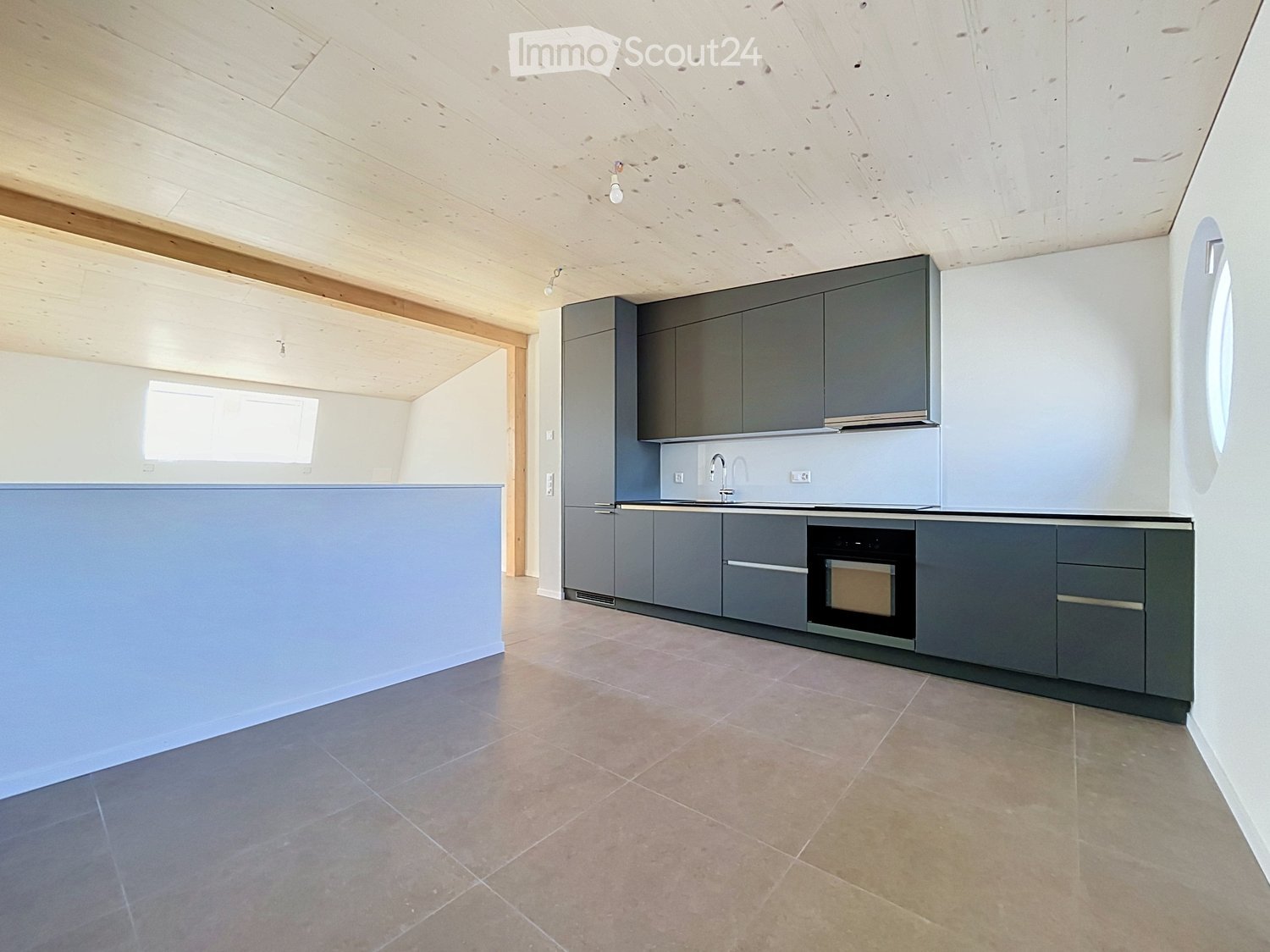 Modern kitchen with grey cabinets, a central island, and a built-in oven. The space features light wooden beams on the ceiling and large windows allowing natural light.