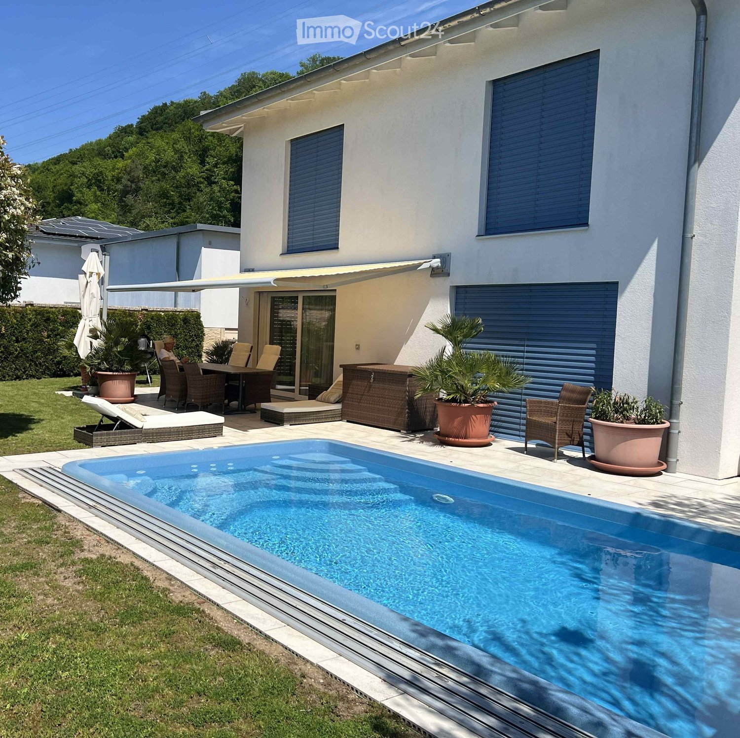 Modern outdoor area with a swimming pool, patio furniture, lounge chairs, and potted plants near a house with large windows and a covered patio.