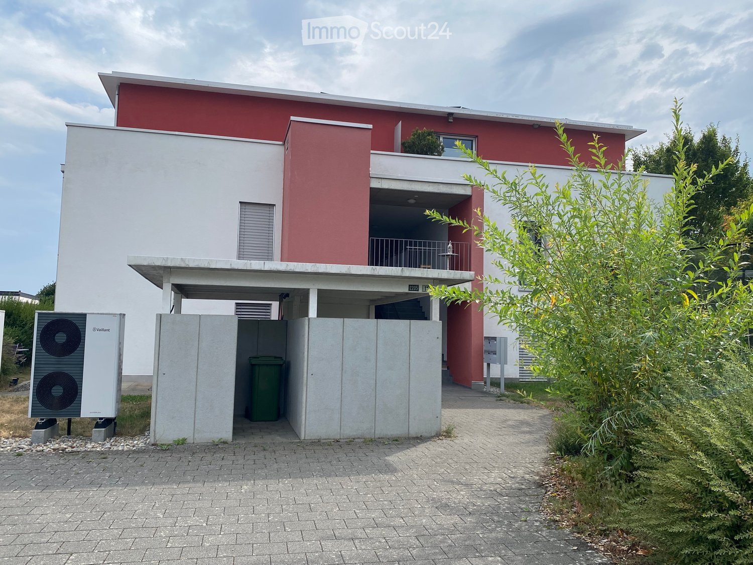 Modern two-story house with red and white exterior, balcony on the upper level, and a covered parking area with a trash bin.