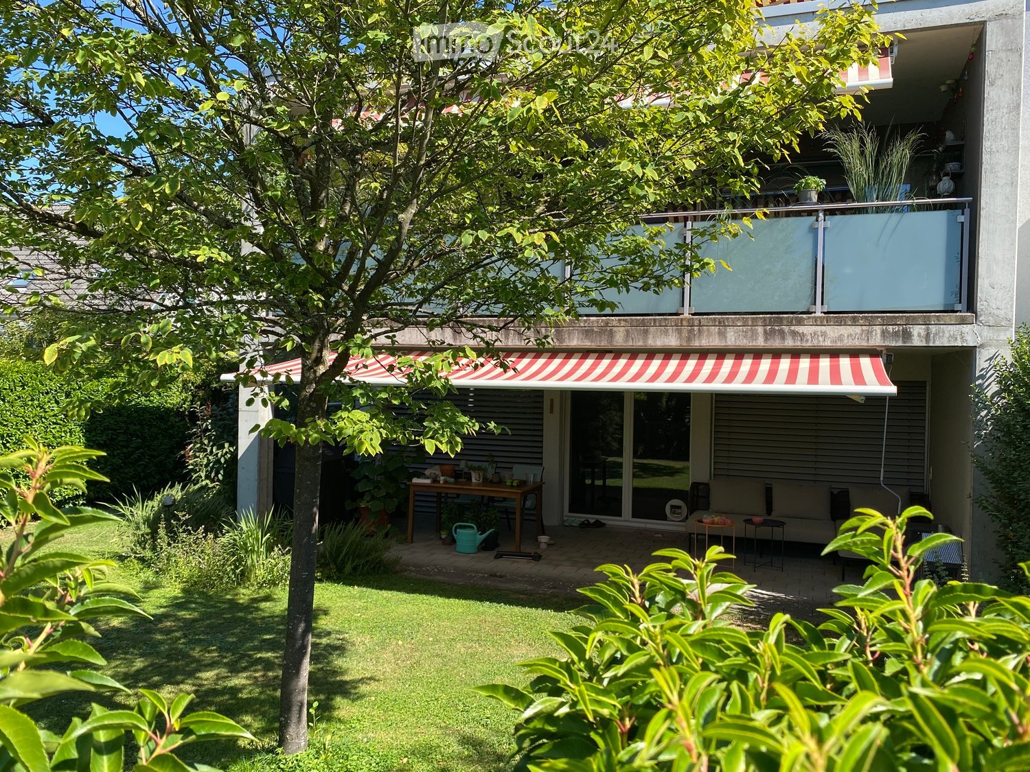 Garden with a grassy lawn, a tree, a patio area with a table, chairs, a sofa, and a small awning. There is also a balcony above with plants and furniture.