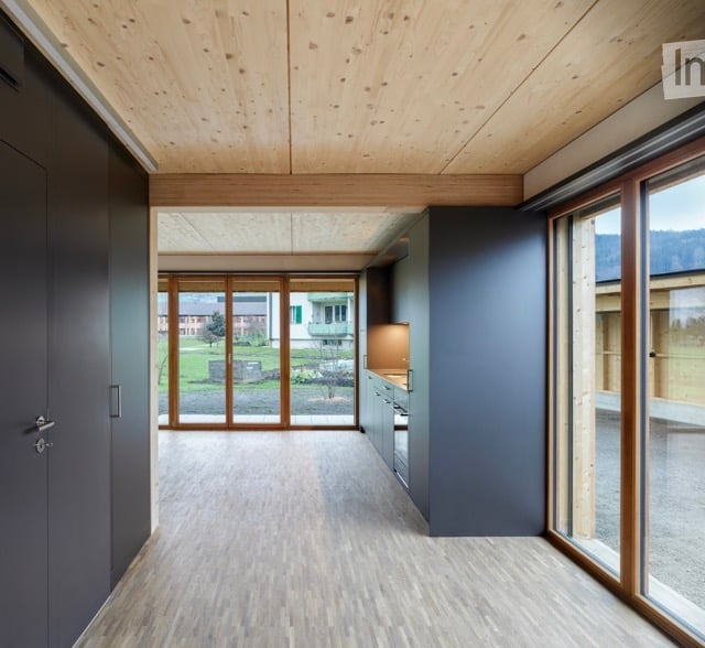 Open-plan unfurnished space with wooden ceiling and floor, large glass doors leading to outdoor view, grey walls, and minimal built-in cabinetry.