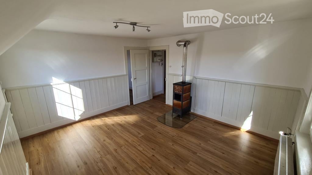 Empty room with wooden flooring, white walls, and a fireplace in the corner. Natural light streams in from a window on the left side.