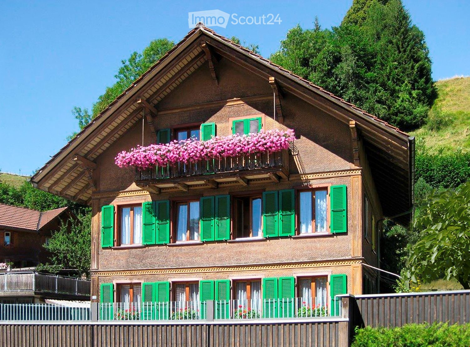 Traditional two-story wooden house with green shutters, multiple windows, and flower boxes on the balcony.