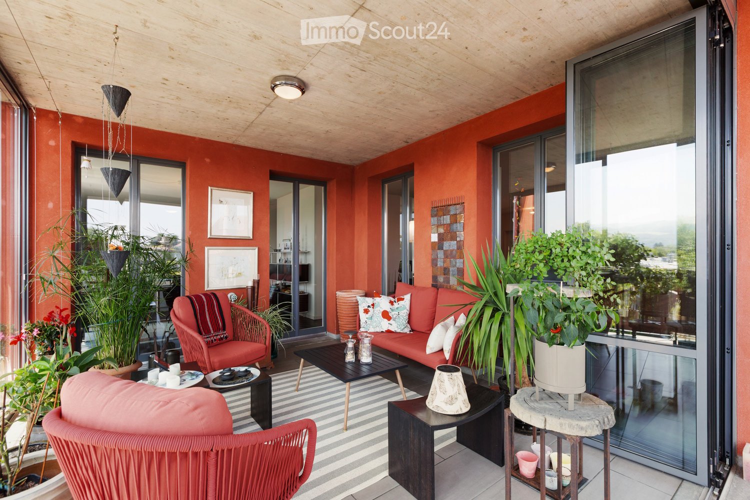 A modern balcony with red walls, wooden ceiling, and large glass doors leading to the interior. Features a red sofa, colorful cushions, a wicker chair, a coffee table, and several potted plants.