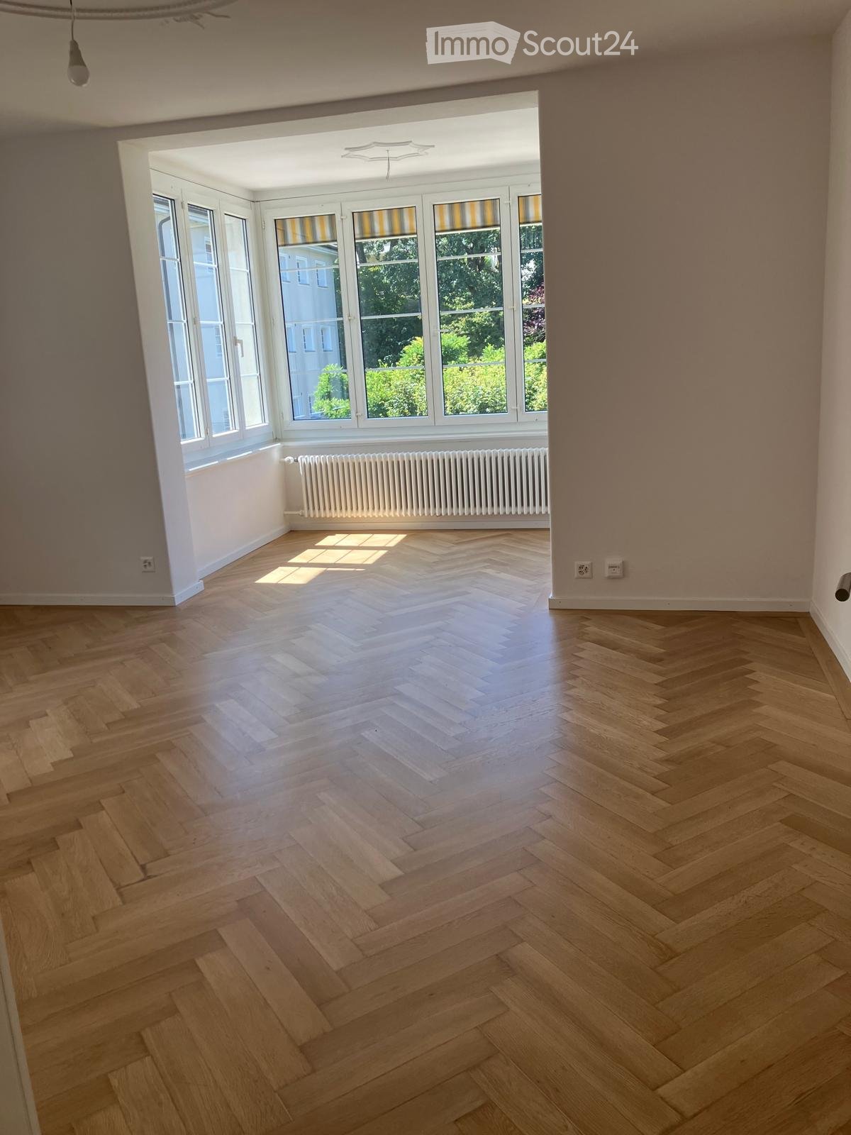 Empty room with light wooden herringbone flooring, large windows with striped blinds, radiator, and visible electrical outlets.