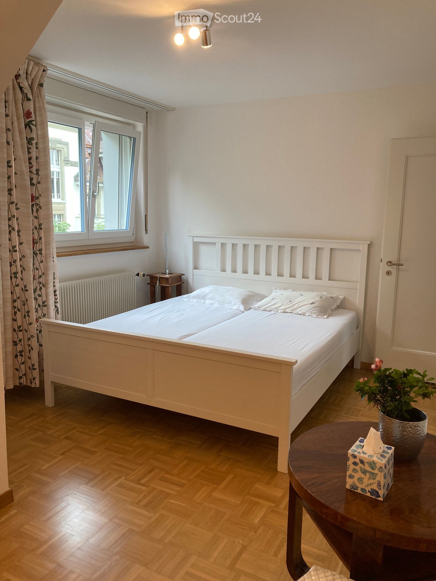 Cozy bedroom with a white wooden bed, nightstand, small round table with a plant, and a tissue box. The room features wooden flooring and large windows with floral curtains.