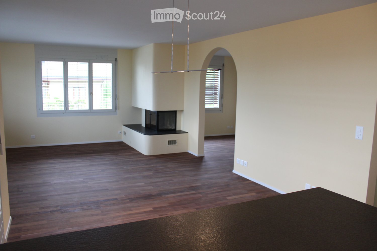 Empty room with wooden flooring, light-colored walls, and a built-in fireplace. Large windows provide natural light, and the space is unfurnished.
