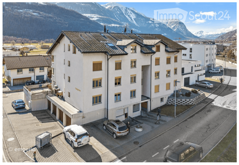 Multi-story residential building with white facade, yellow window frames, and a tiled roof, situated in a scenic mountainous area with a parking lot and surrounding greenery.