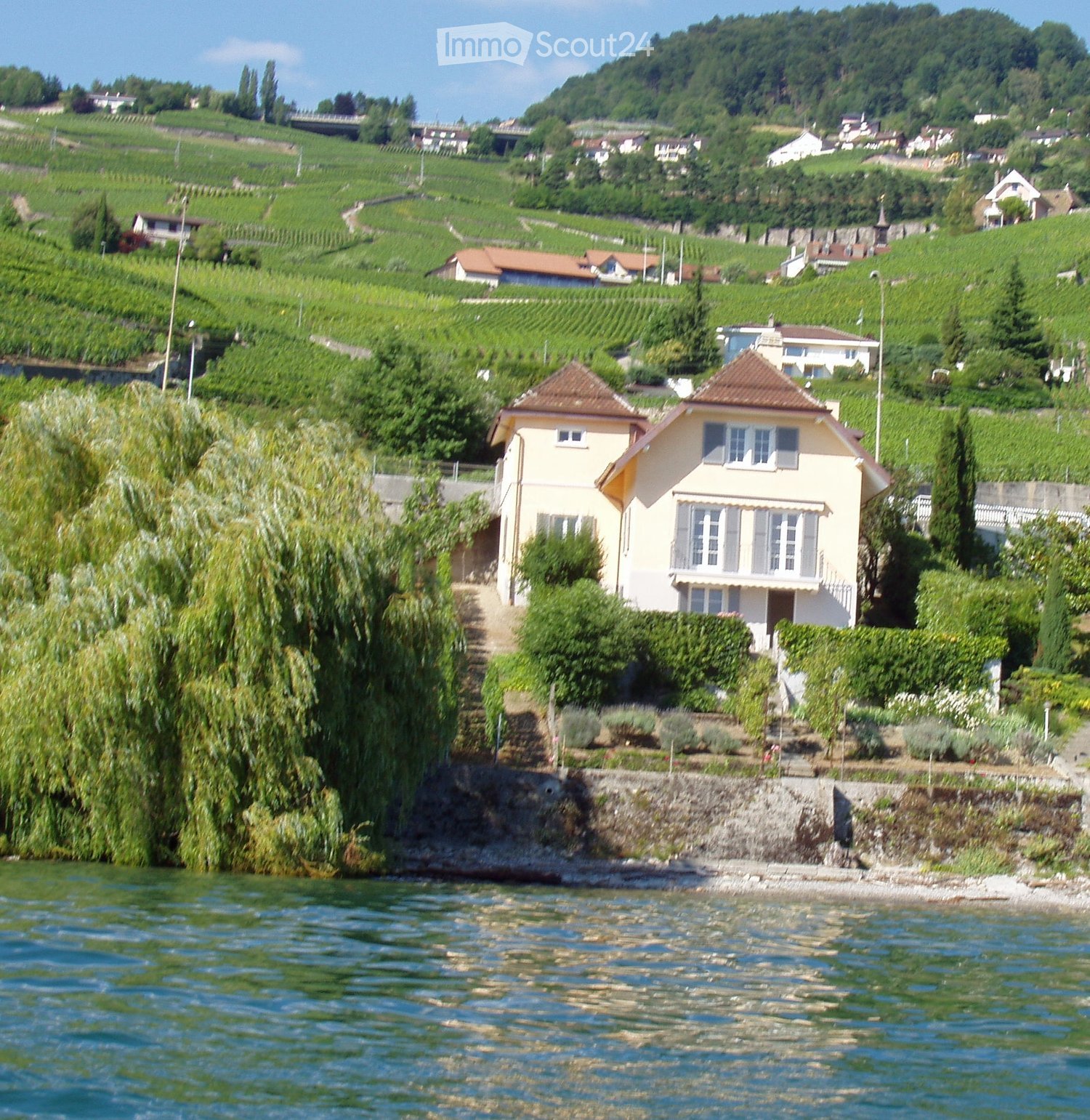 A two-story house with a beige exterior and a red-tiled roof, situated on a hillside overlooking a body of water, surrounded by greenery and vineyards.