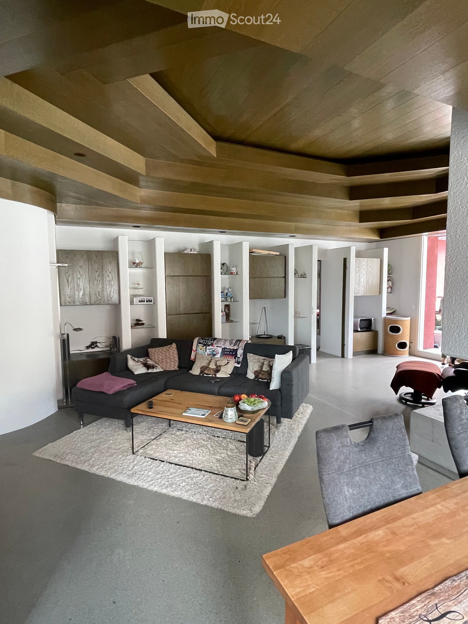 Modern living room with a grey sectional sofa, wooden coffee table, plush rug, and a dining area with a wooden table and chairs. The room features built-in cabinets and a sleek, contemporary design.