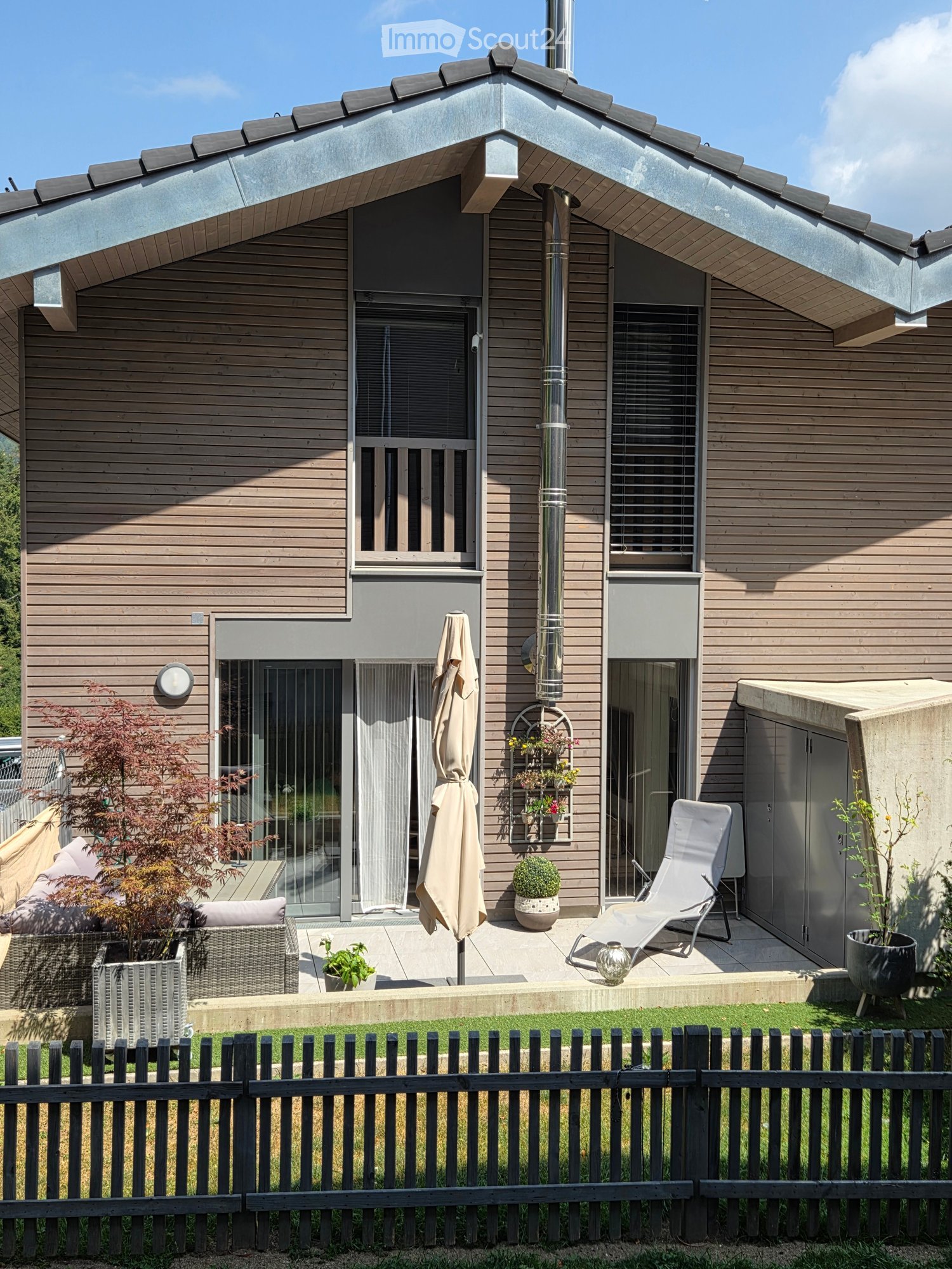 Modern wooden house with a patio area featuring a wicker chair, umbrella, sun lounger, and potted plants.