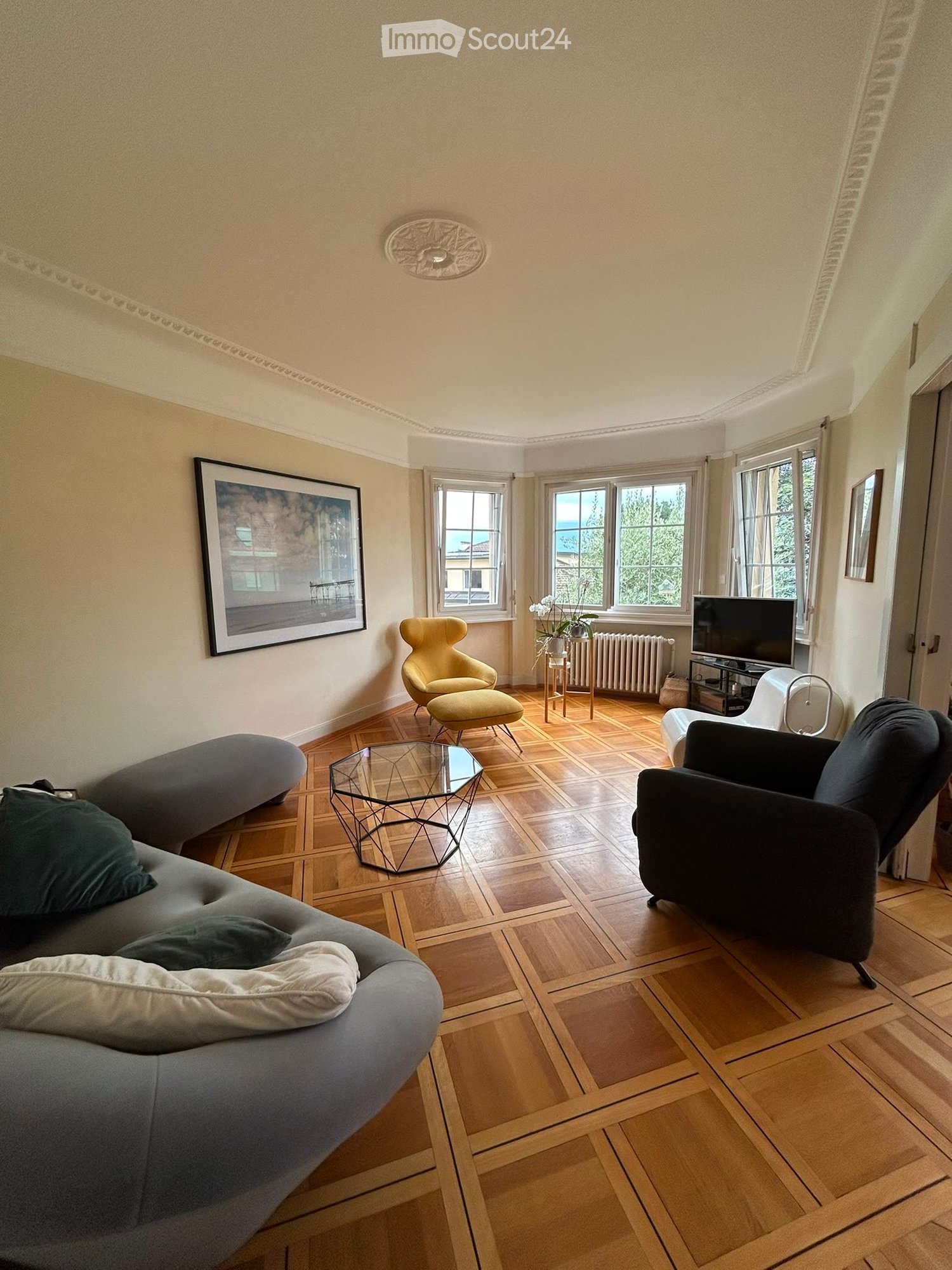Living room with wooden flooring, large windows, a grey sofa, a yellow armchair, a glass coffee table, and a television on a stand.