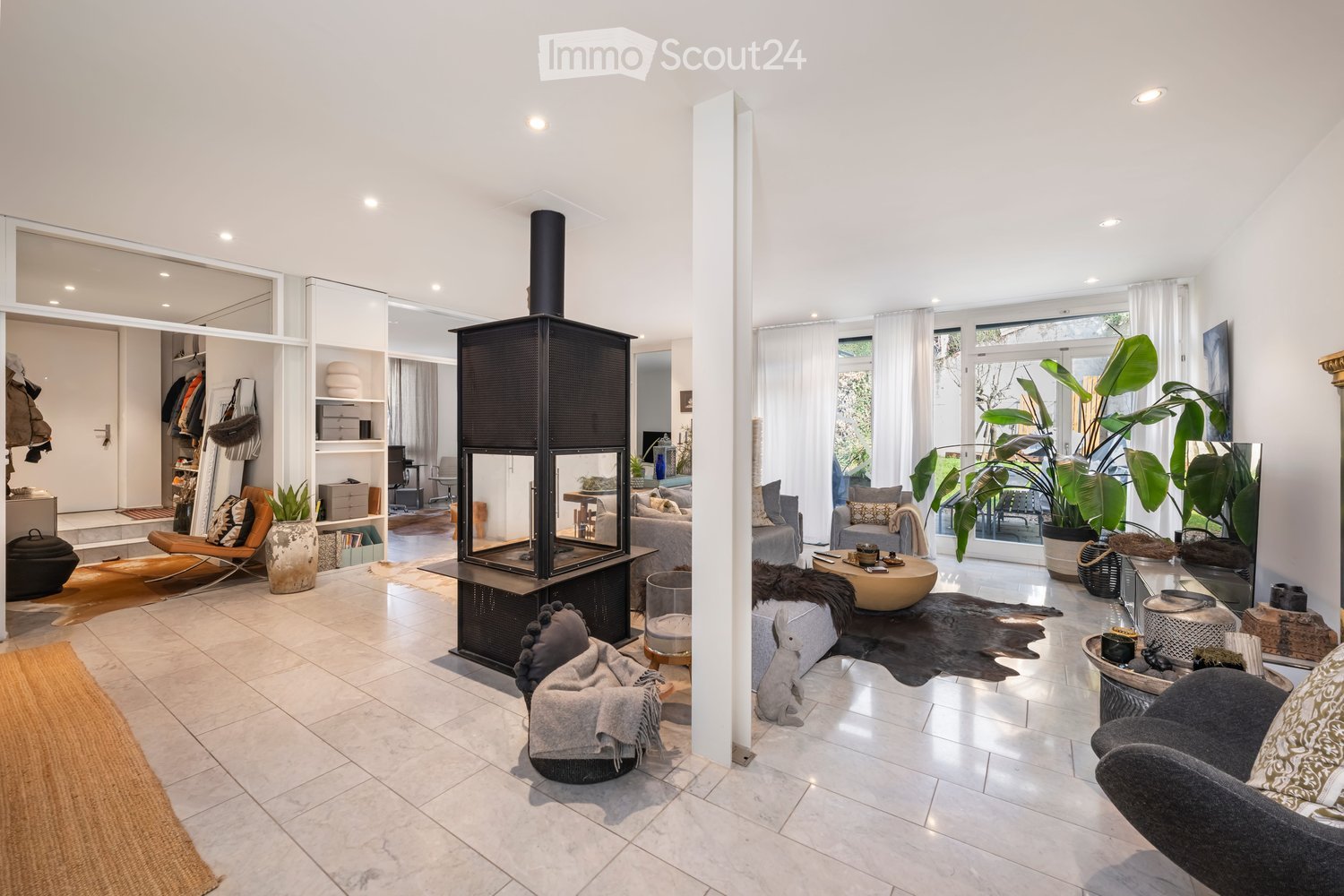 Modern living room with a fireplace, large windows, and contemporary furniture including a sofa, coffee table, armchair, and decorative plants.