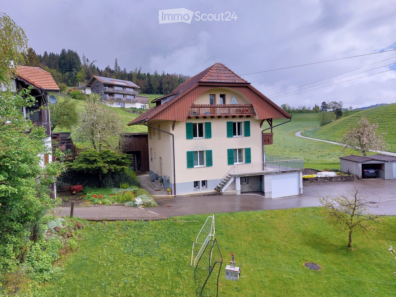 Two-story house with red roof, green shutters, and a balcony. The house is surrounded by a well-maintained lawn, garden, and a driveway leading to a garage. The property is situated on a hillside with a scenic view of the surrounding landscape.