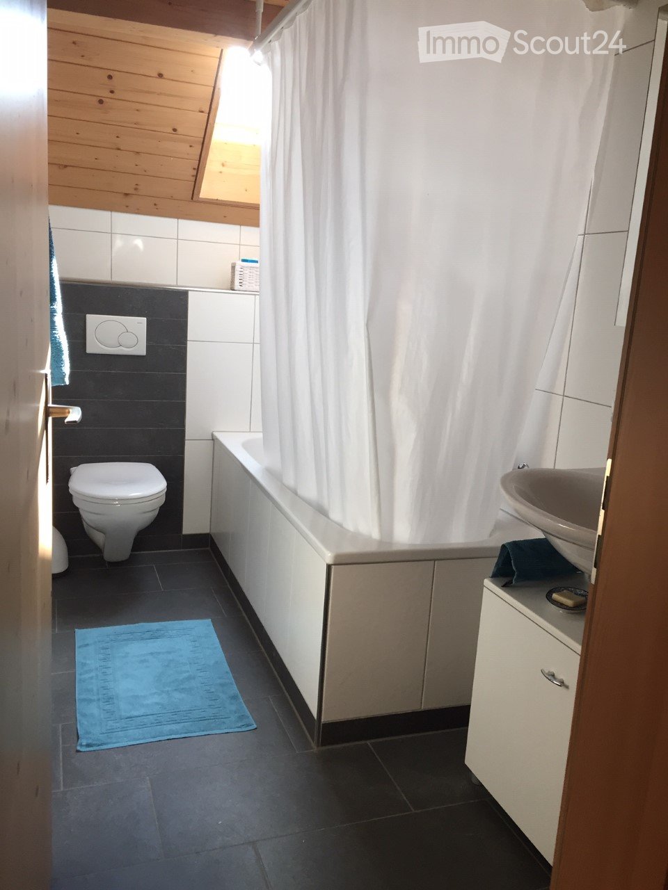 Modern bathroom with dark tiles, white fixtures, a bathtub, toilet, sink, and a blue bath mat.