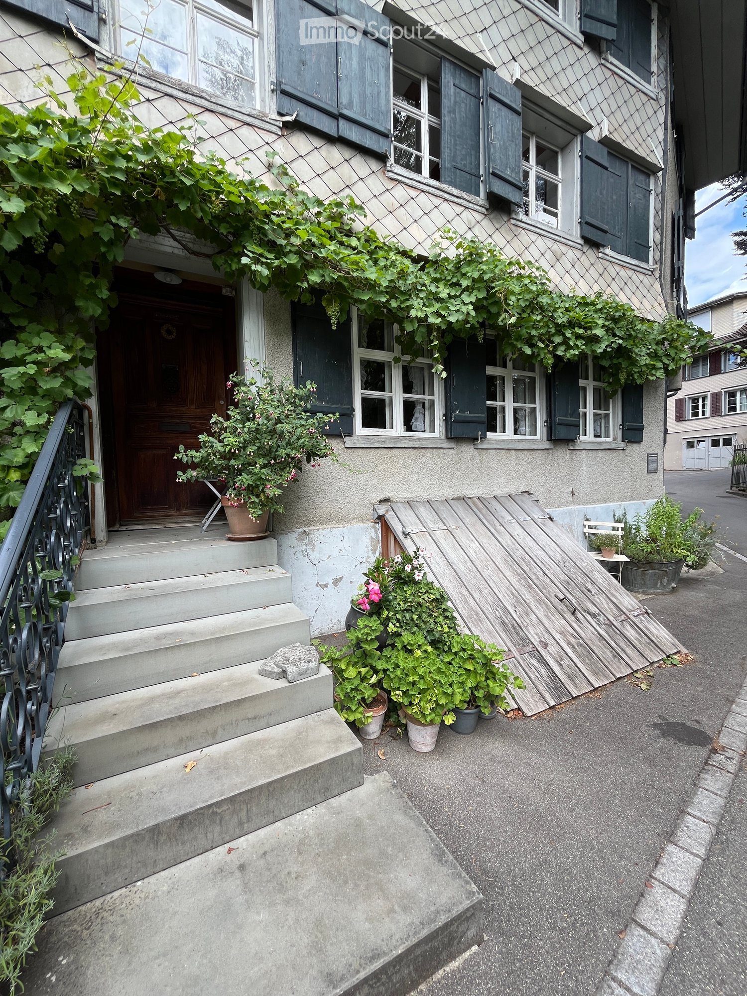 A two-story building with a stone facade, multiple windows with shutters, and a wooden ramp leading to the entrance. The entrance has a wooden door, steps, and potted plants on either side.