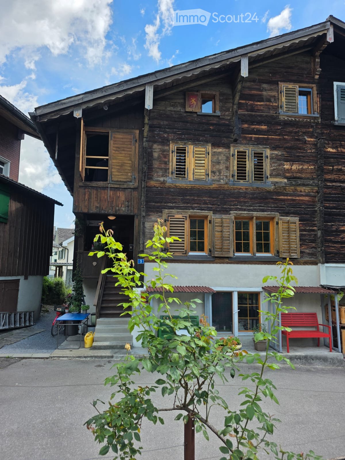 Traditional wooden house with multiple windows, shutters, and a small porch area with a red bench and plants.