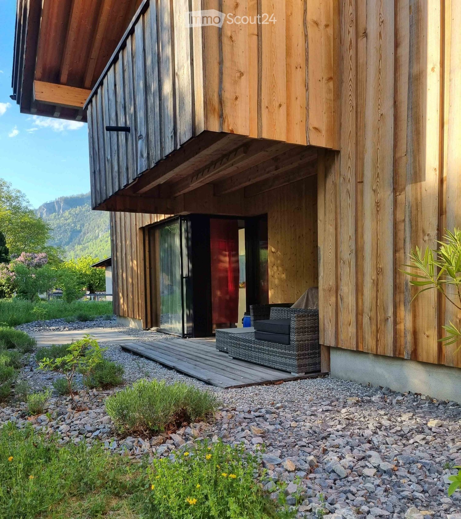 Wooden house exterior with a covered entrance, a wicker chair and table on a wooden deck, surrounded by gravel and greenery.