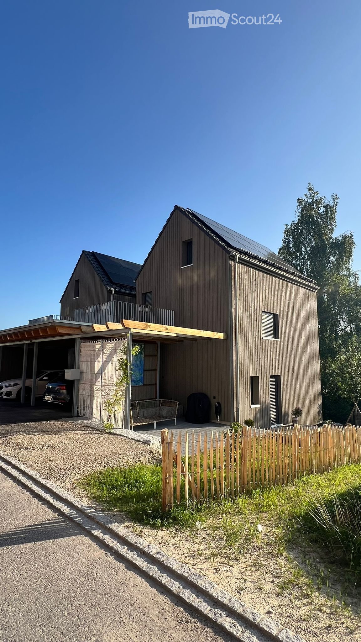 Modern two-story house with wooden siding, solar panels on the roof, a covered garage, and a small outdoor seating area.