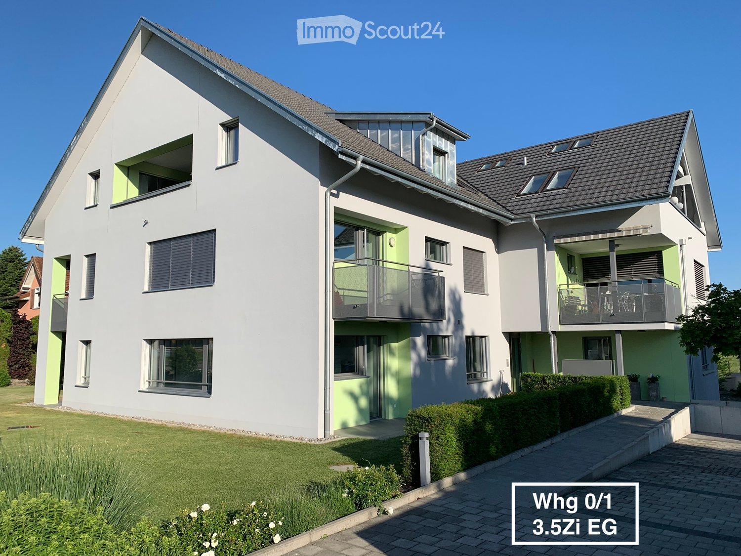 Modern two-story house with white and green exterior, multiple windows, balconies, and a well-maintained lawn.