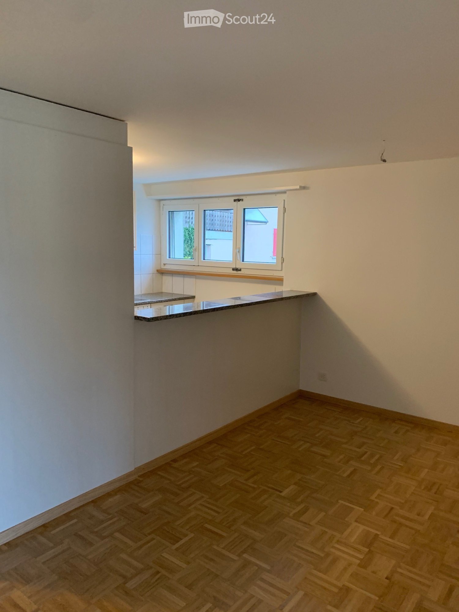 Empty room with wooden herringbone flooring, white walls, and a window with white frame and blinds. There is a small countertop with a sink and a window above it.