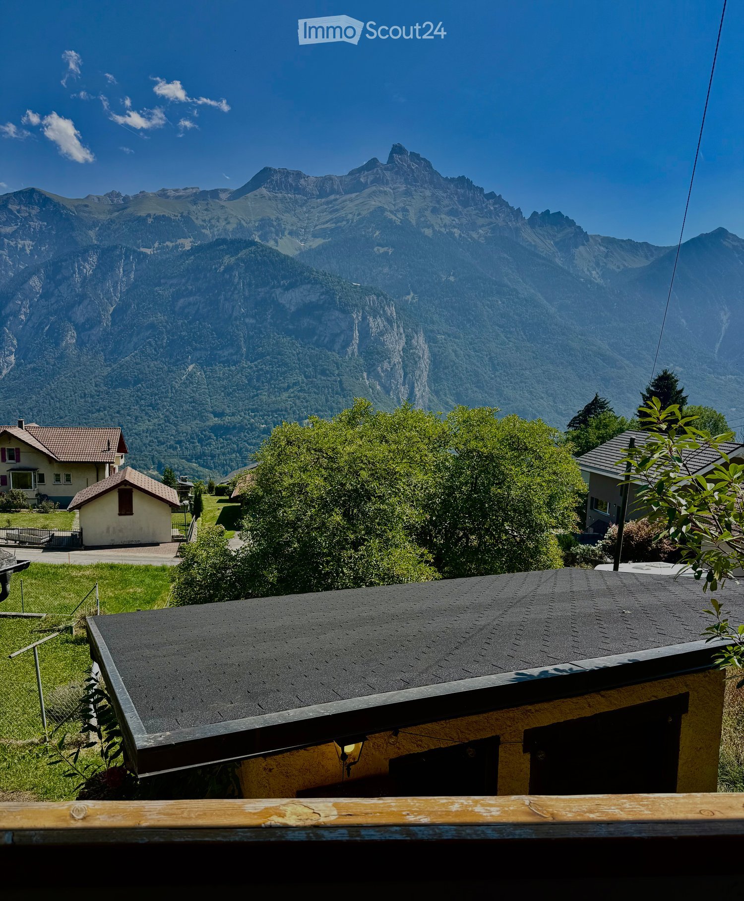 A serene outdoor view from a rooftop, showcasing a lush green garden, several houses, and a majestic mountain range under a clear blue sky.