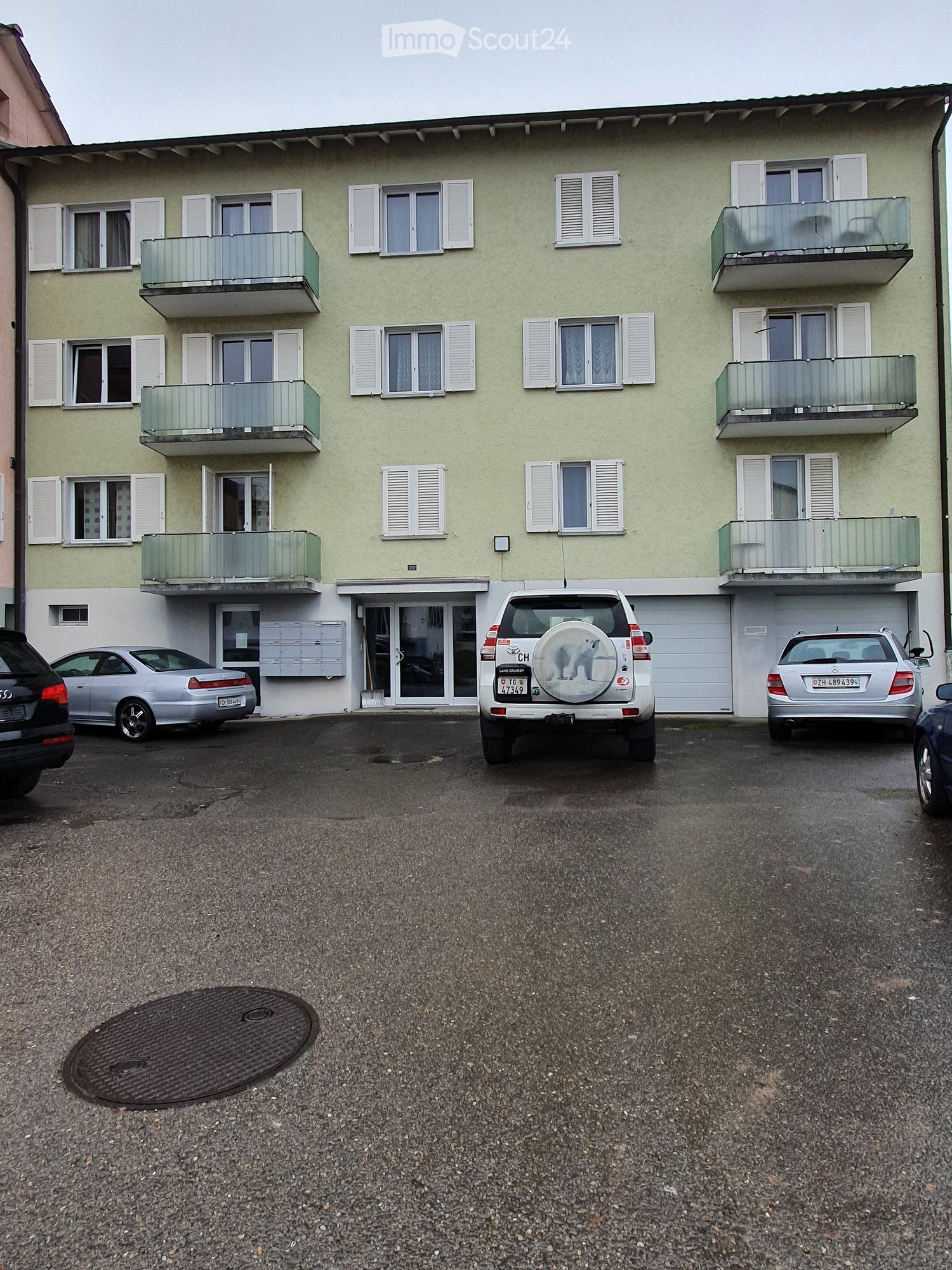 A three-story residential building with light green walls, white shutters, and multiple balconies. Several cars are parked in front of the building, and there is a manhole cover on the ground.