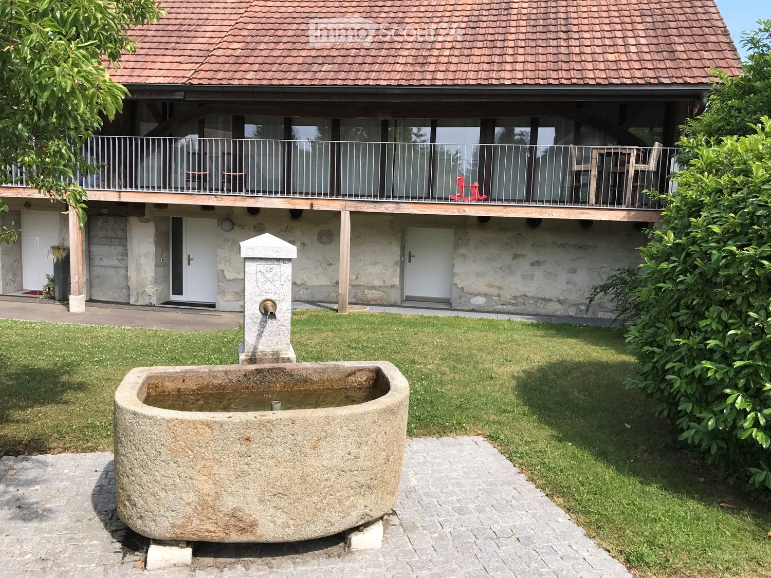 A two-story house with a balcony, stone walls, and a well-maintained garden featuring a stone water basin and a fountain.