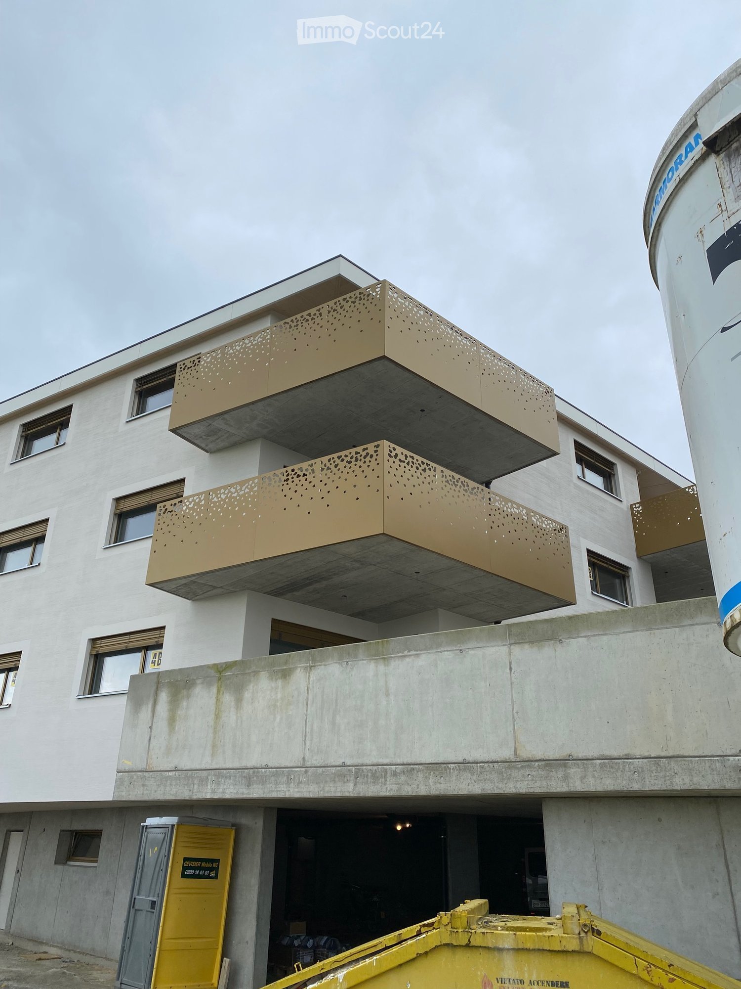 Modern multi-story building with white facade, large windows, and distinctive gold-colored balconies. Features a raised ground floor and a visible garage entrance.