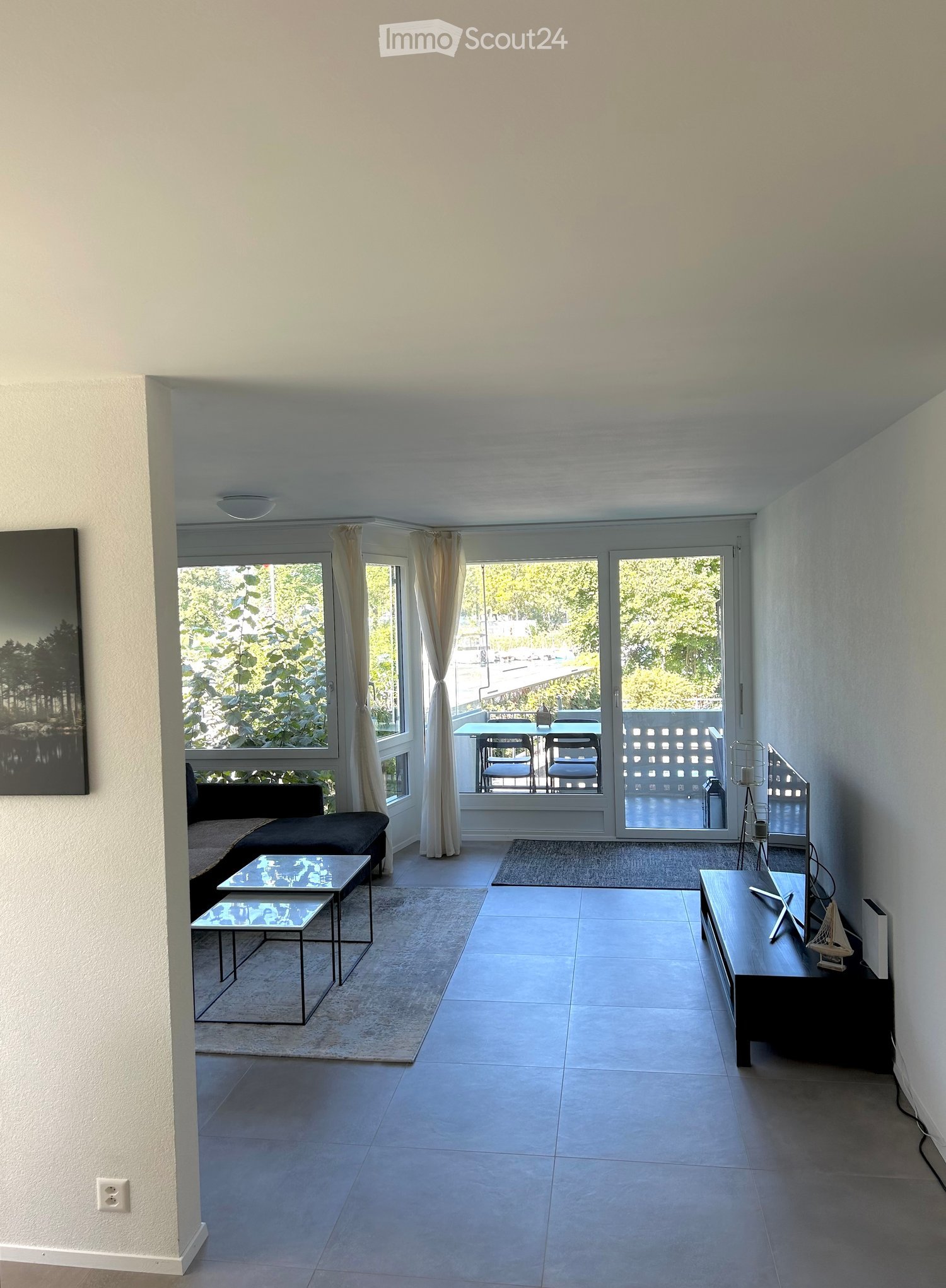 Modern living room with large windows and a balcony, featuring a sofa, coffee table, TV stand, and chairs.