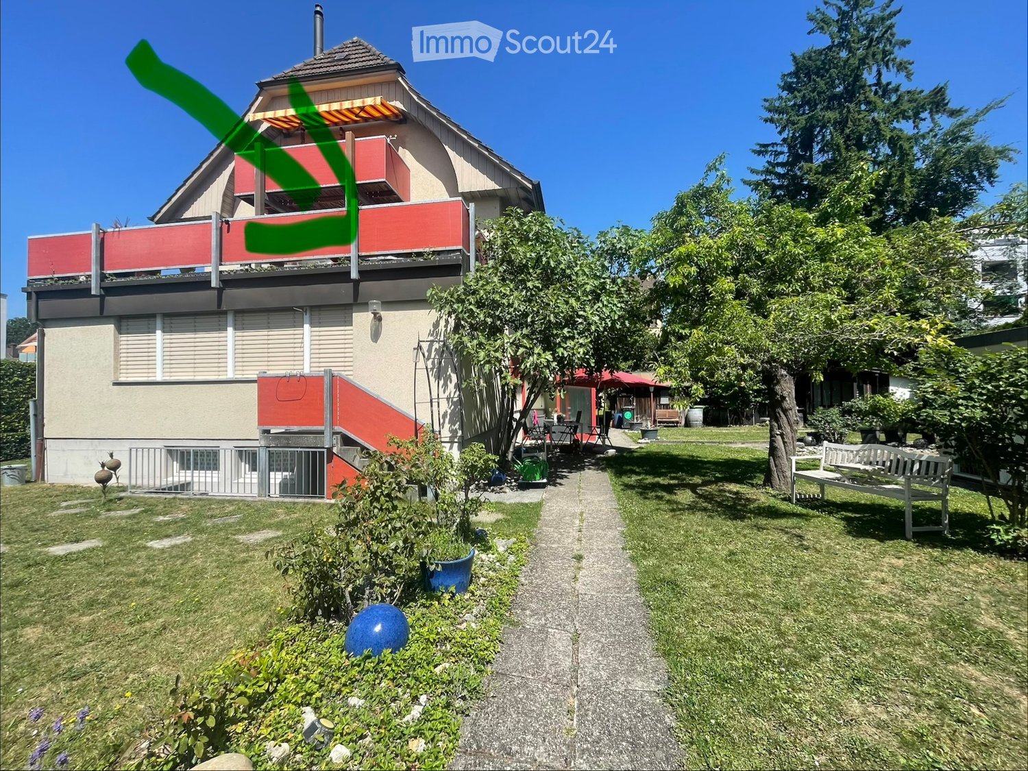 A two-story house with a red balcony and a well-maintained garden featuring a bench, pathway, and various plants.