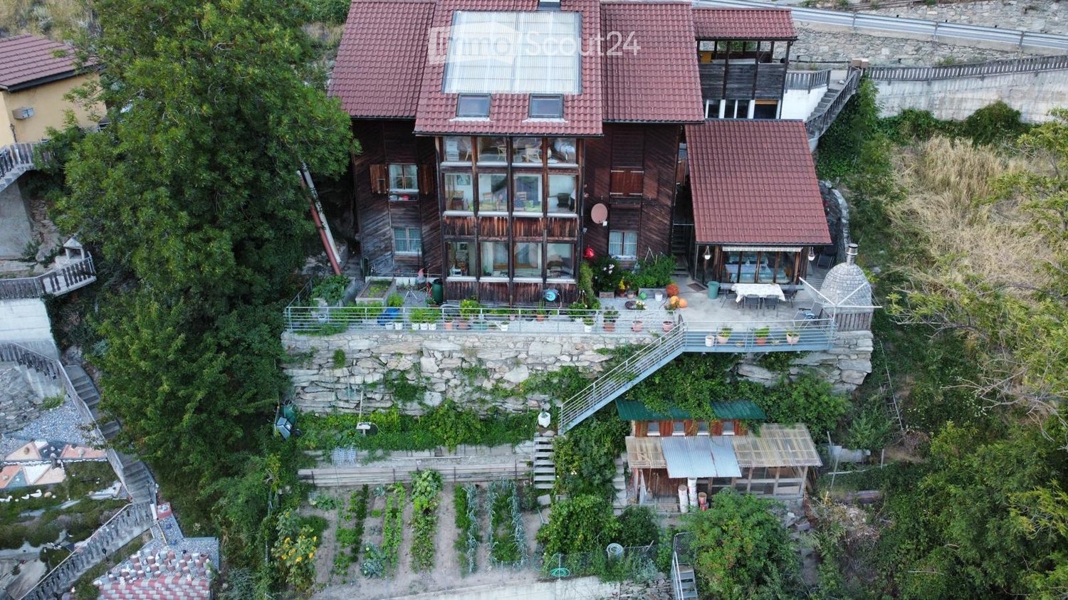 A multi-level wooden house with red-tiled roofs, situated on a raised stone foundation surrounded by greenery and a small garden area.