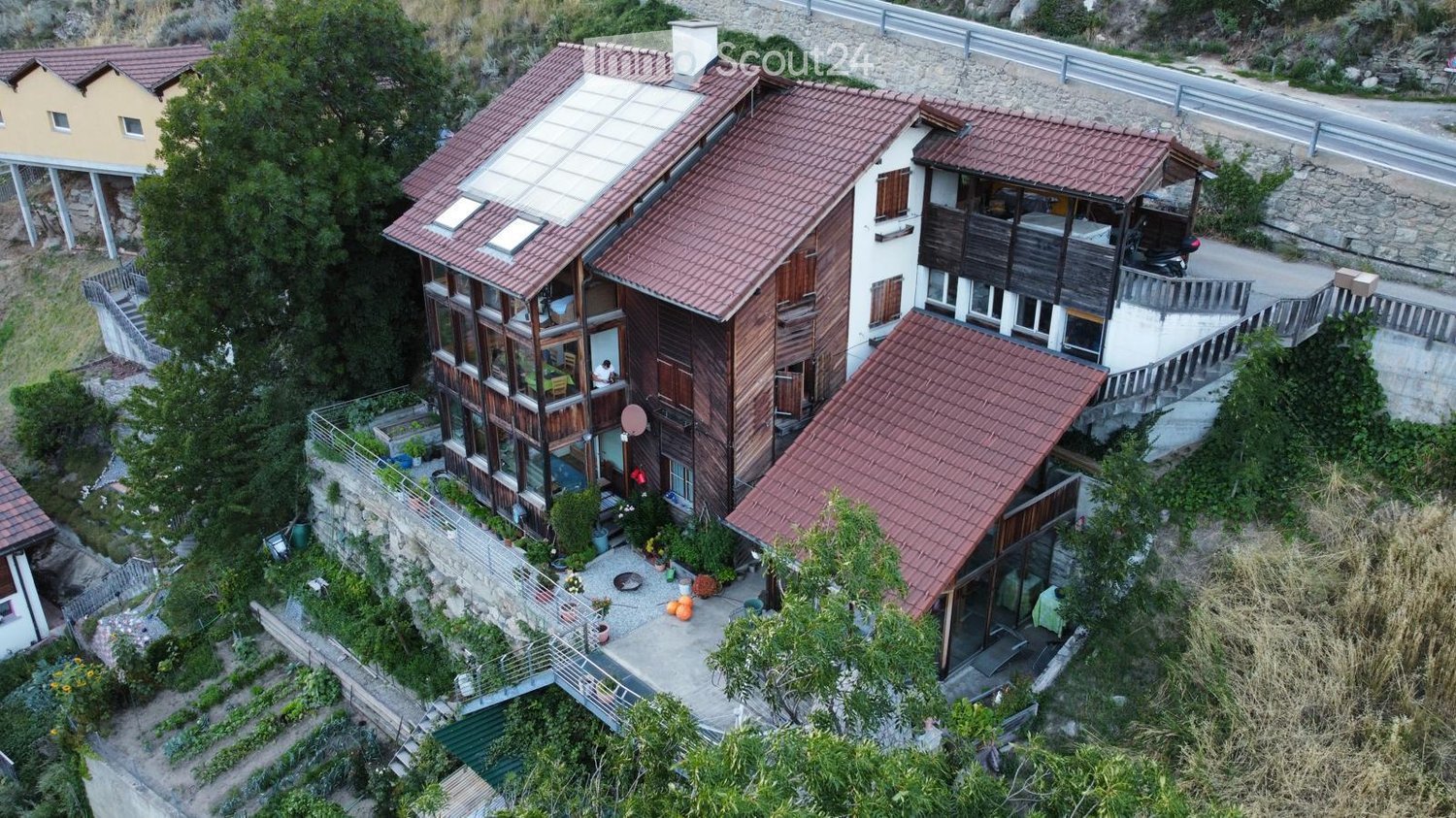 A multi-story residential building with red-tiled roofs, wooden balconies, and a garden area. The structure includes multiple levels with large windows and outdoor spaces.