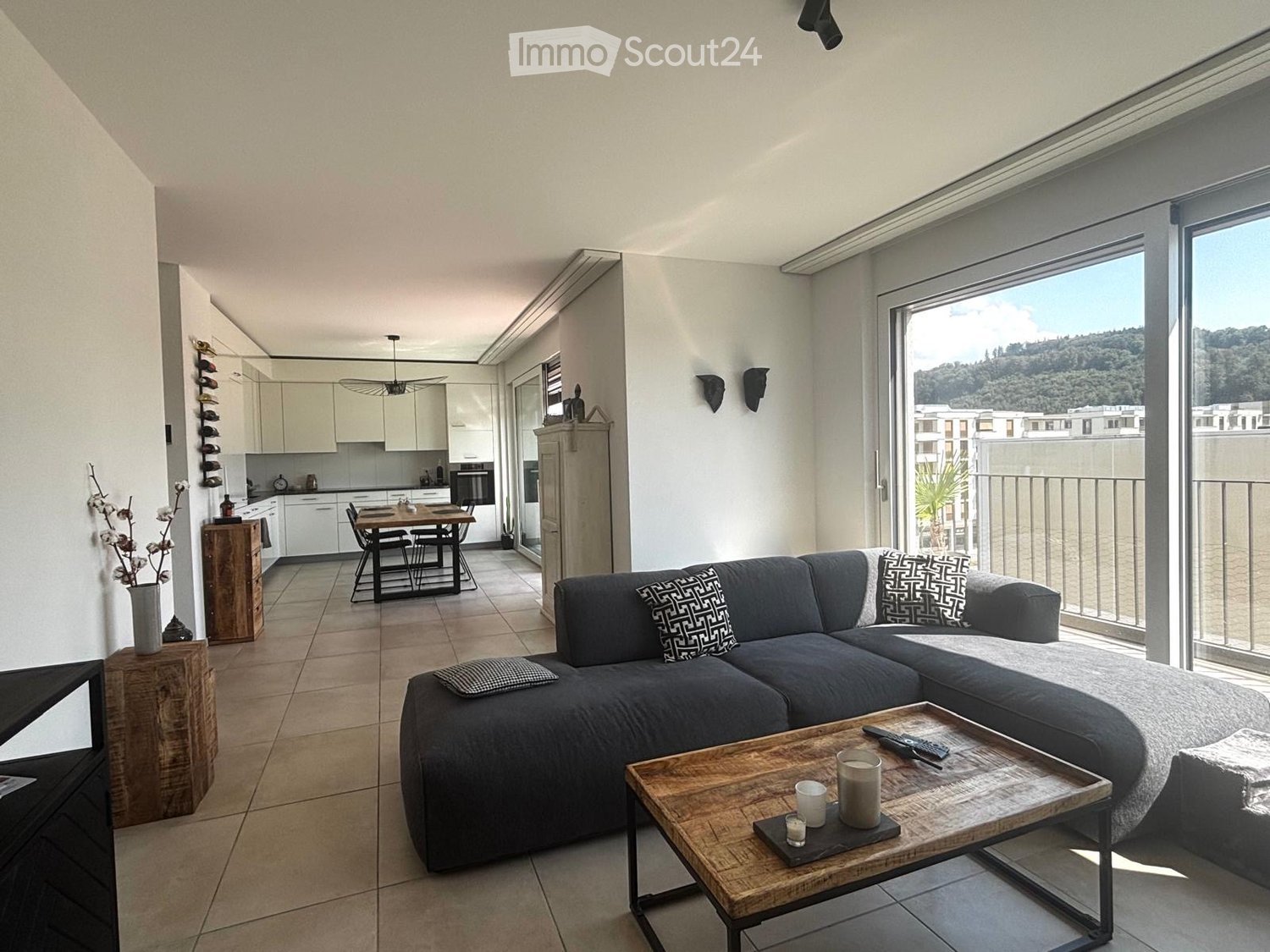 Modern living room with grey sectional sofa, wooden coffee table, dining area with chairs and table, kitchen area with cabinets and appliances, and large windows offering a scenic view.