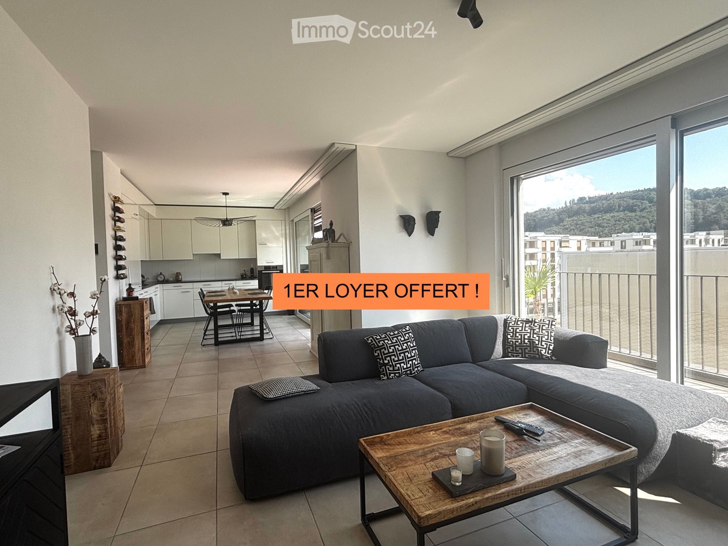 Modern living room with a grey sectional sofa, wooden coffee table, dining area, and large windows offering a scenic view.