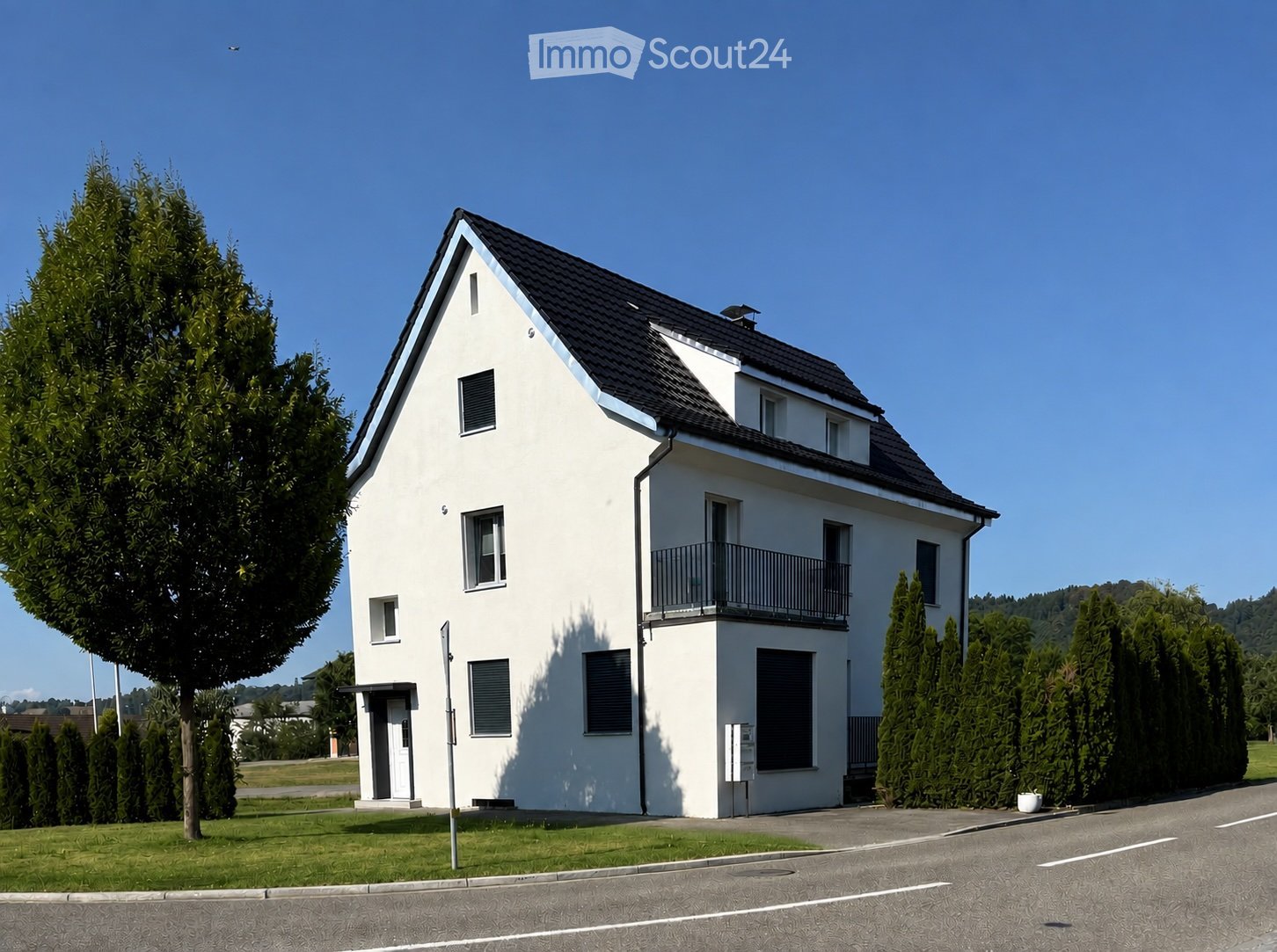 Modern two-story white house with a dark roof, multiple windows, and a small balcony on the upper level. The house is surrounded by well-maintained greenery and a paved driveway.