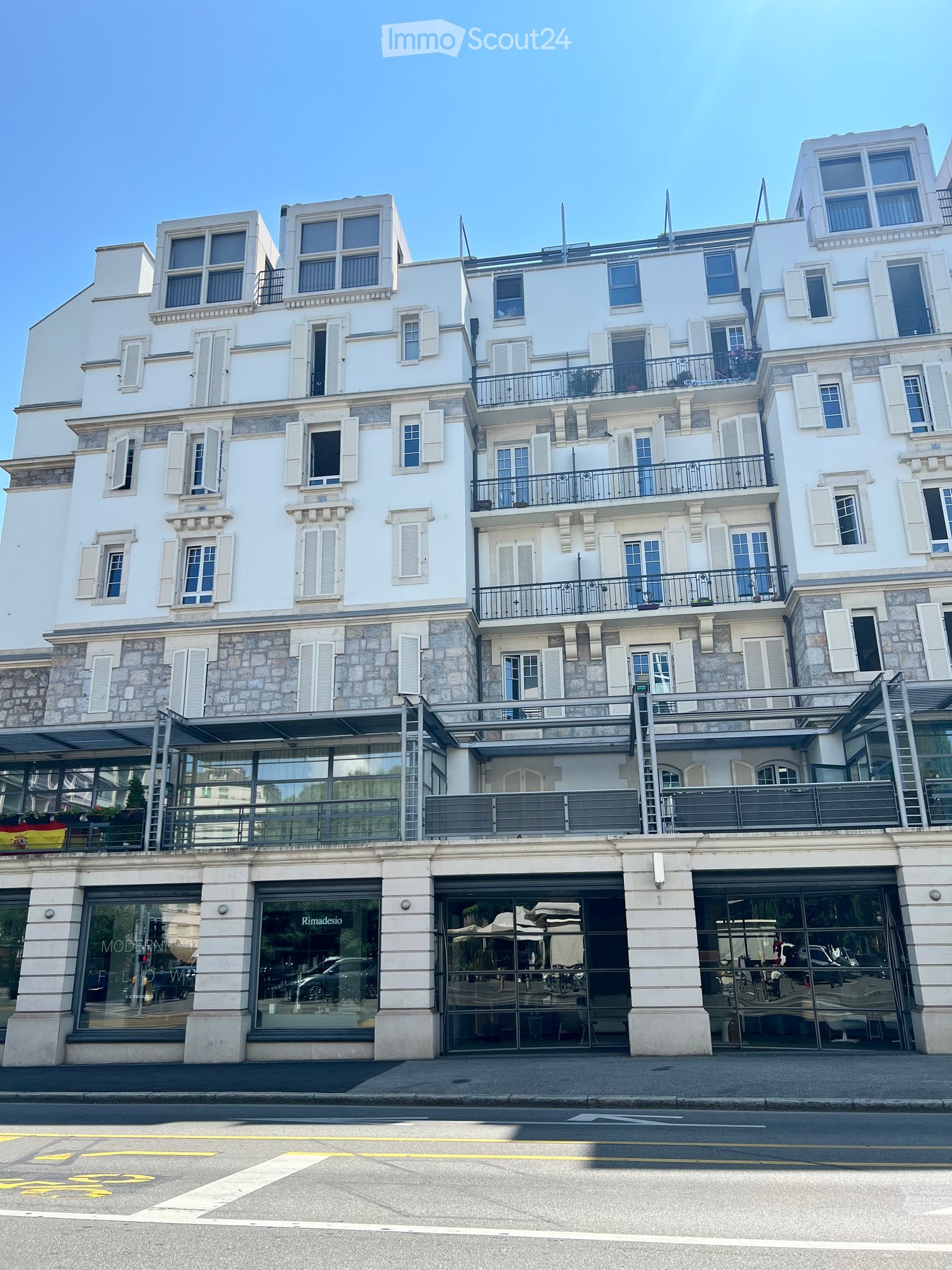A multi-story white building with stone accents, multiple windows, balconies, and a modern storefront with glass doors and signage.
