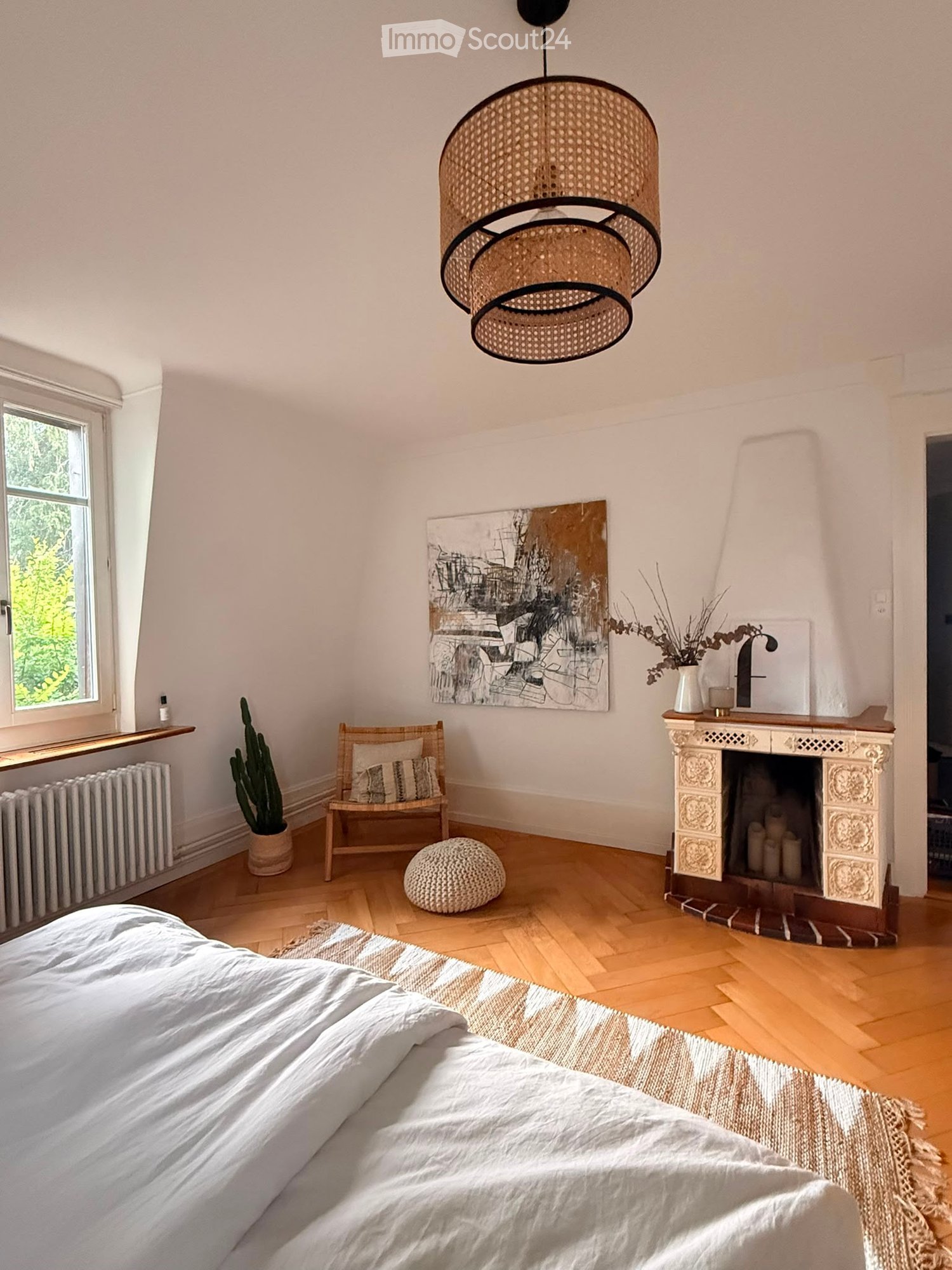 Bright bedroom with wooden flooring, a large bed with white bedding, a fireplace with candles, a wooden chair with a cushion, a round woven stool, a cactus in a pot, a painting on the wall, and a hanging woven lamp.
