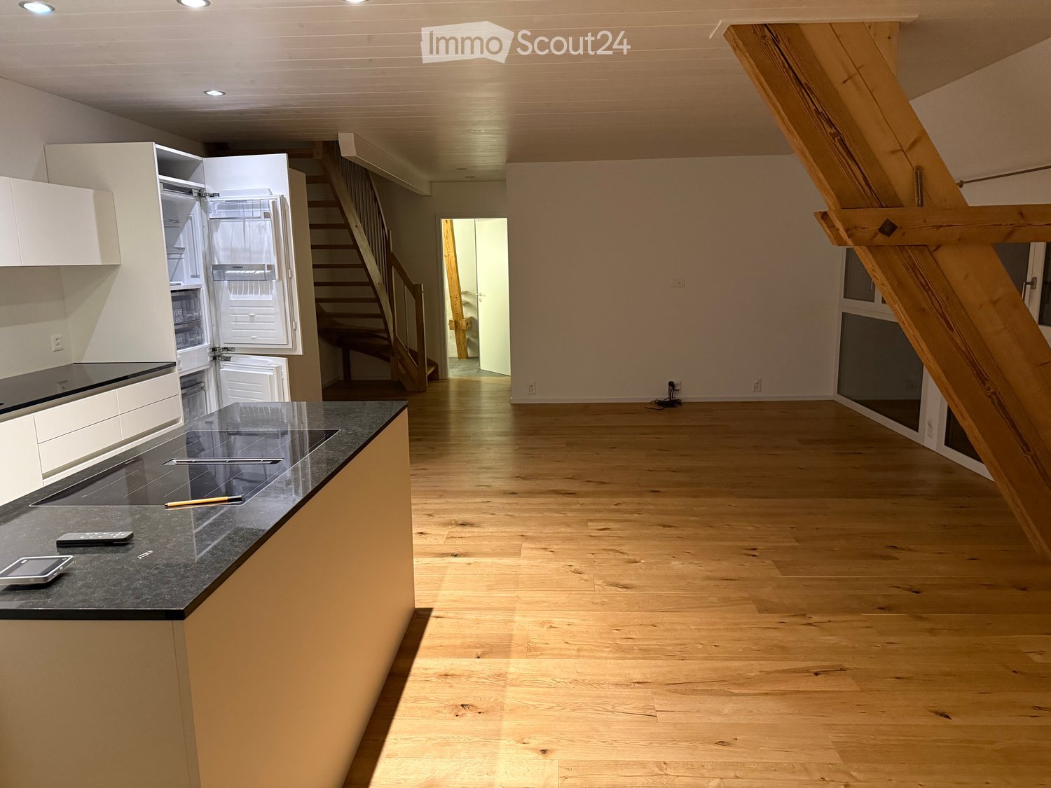 Open-plan kitchen and living area with light wooden flooring, white cabinets, a black countertop, and a staircase leading to another level.