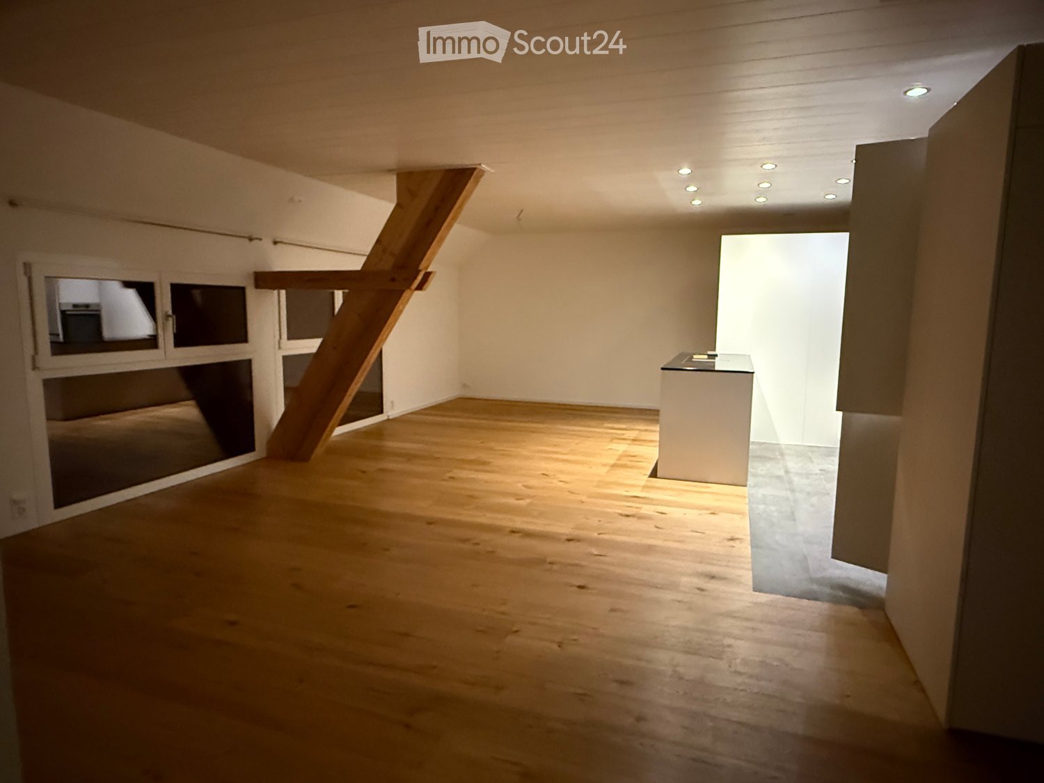 Empty room with wooden flooring, white walls, and recessed ceiling lights. A small white counter is visible in the corner, and a large window with blinds is on the left side.