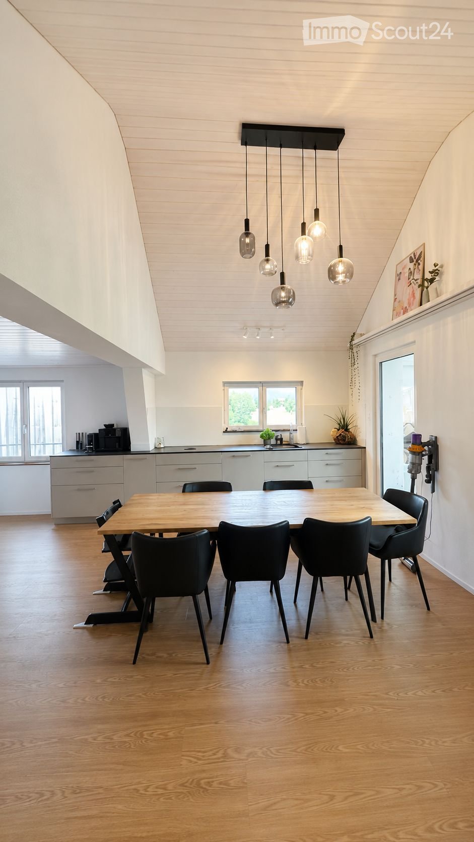 Modern dining area with a wooden table, black chairs, pendant lights, and kitchen cabinets in the background.