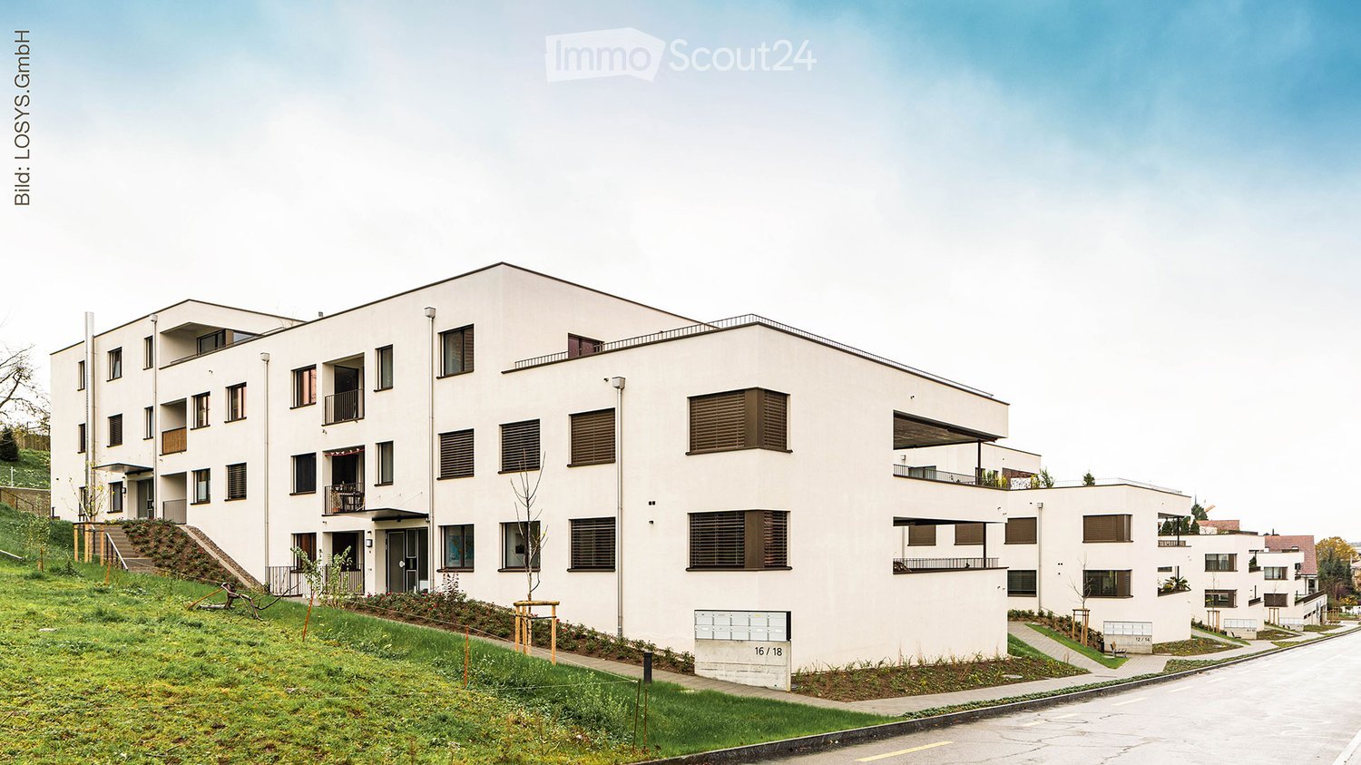 Modern multi-story residential building with white facade, multiple windows, balconies, and surrounding greenery.