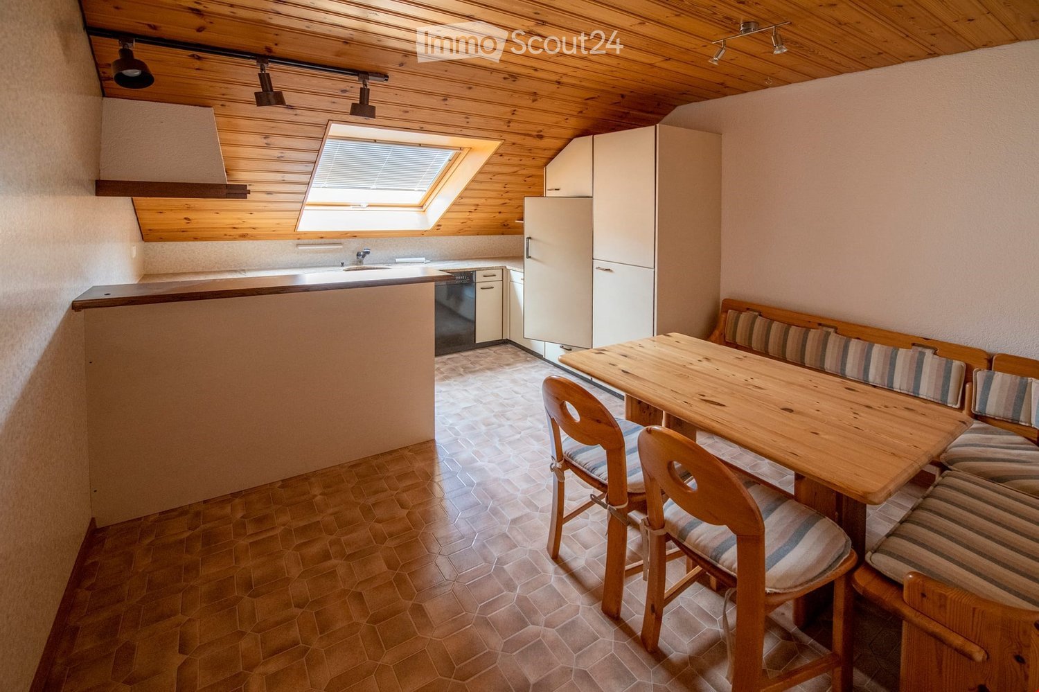 A cozy, well-lit room with a wooden ceiling and exposed beams. It features a kitchen area with a refrigerator, dishwasher, and cabinets, as well as a dining area with a wooden table, chairs, and a bench with striped cushions.