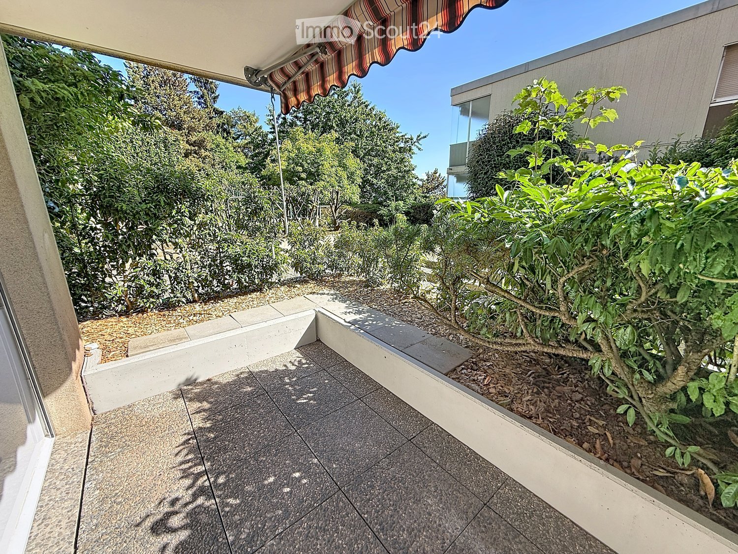 Balcony with tiled floor, small garden area with plants and shrubs, striped awning, and a view of surrounding greenery and buildings.