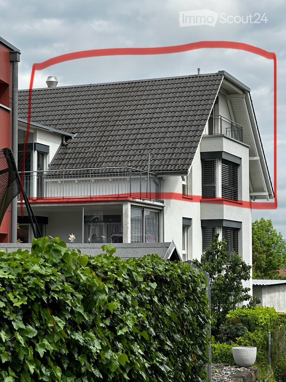 Two-story house with a gray tiled roof, white walls, and a balcony on the upper level. The house features multiple windows and is partially obscured by a dense green hedge in the foreground.